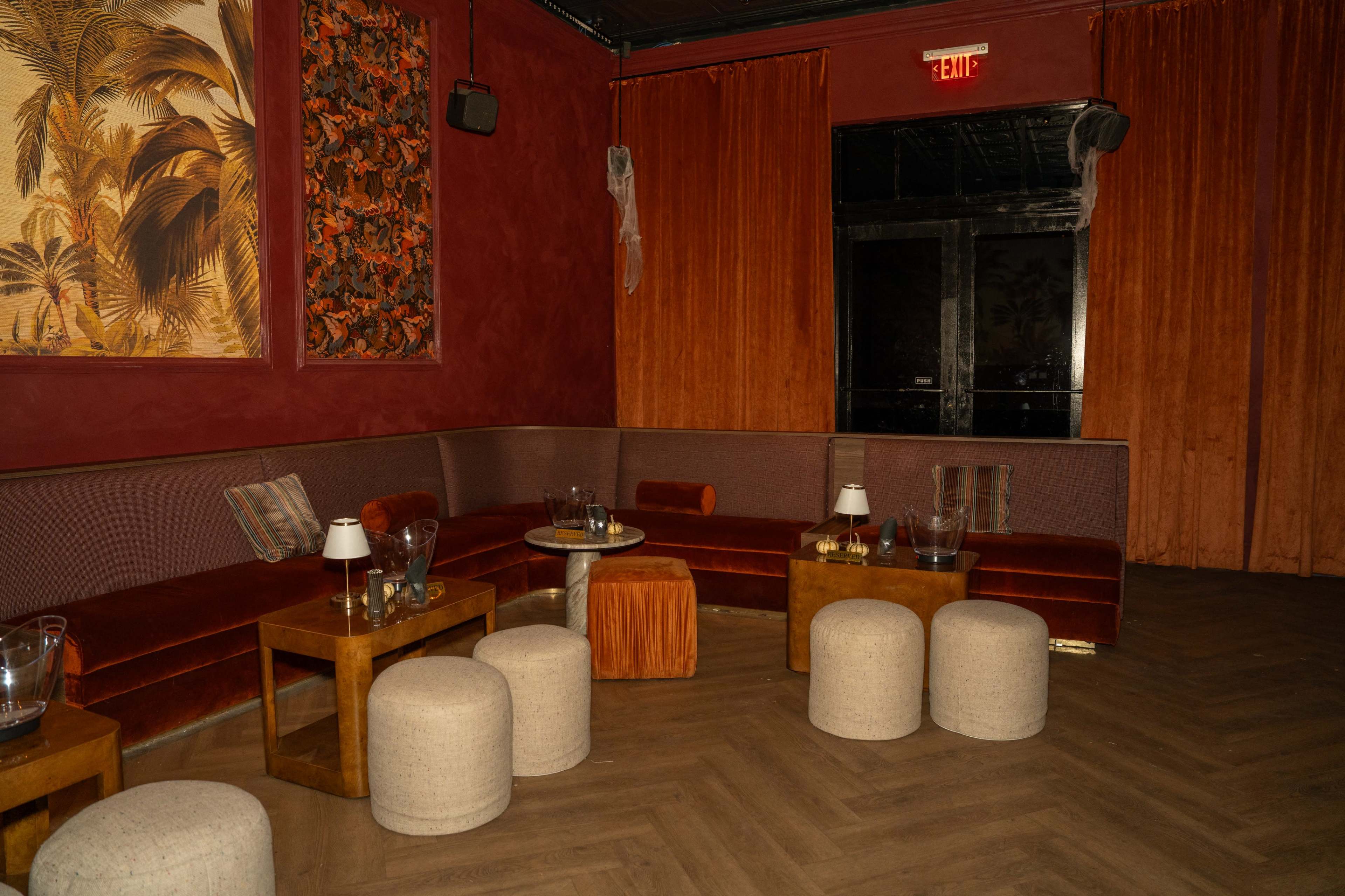The image shows a dimly lit lounge area with red walls, plush seating, wooden furniture, and round stools arranged in a cozy layout.