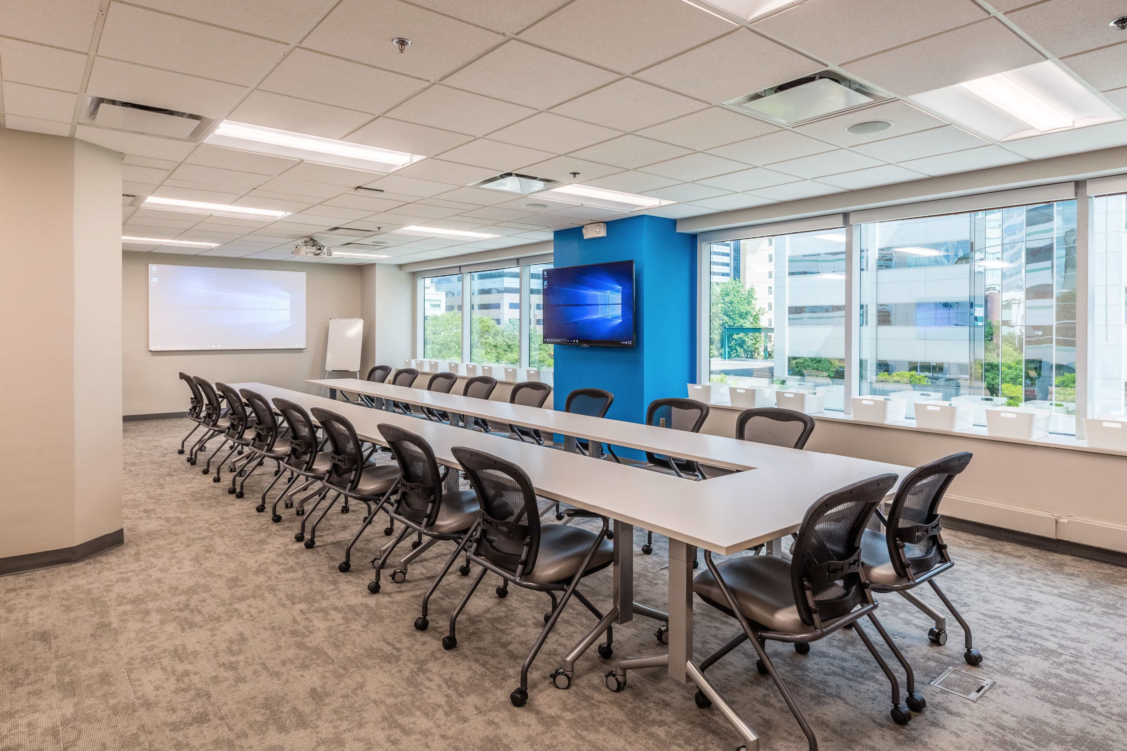 A modern conference room features a long table with rolling chairs, two large screens mounted on the walls, and windows that provide natural light.