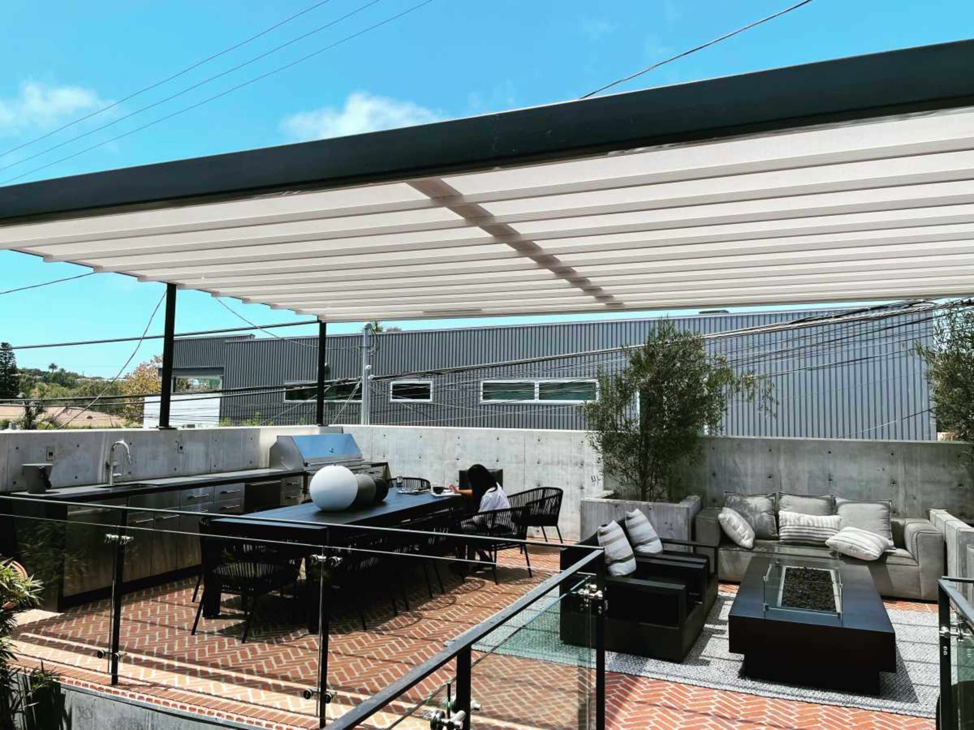 A rooftop terrace features a dining area with a table, sectional sofas, and an outdoor kitchen, surrounded by greenery and buildings in the background.