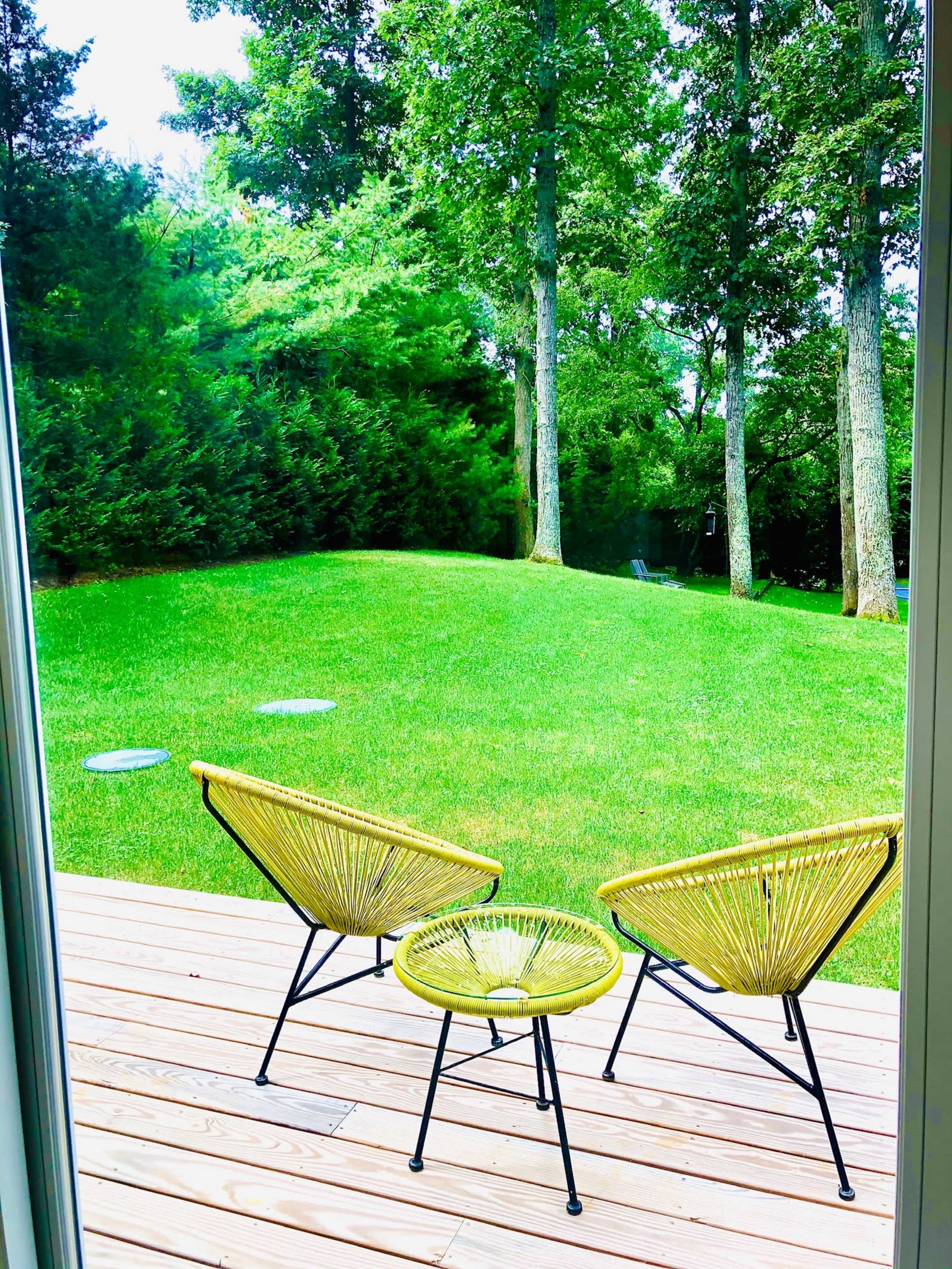 Two yellow chairs and a small table are positioned on a wooden deck overlooking a green lawn and trees.