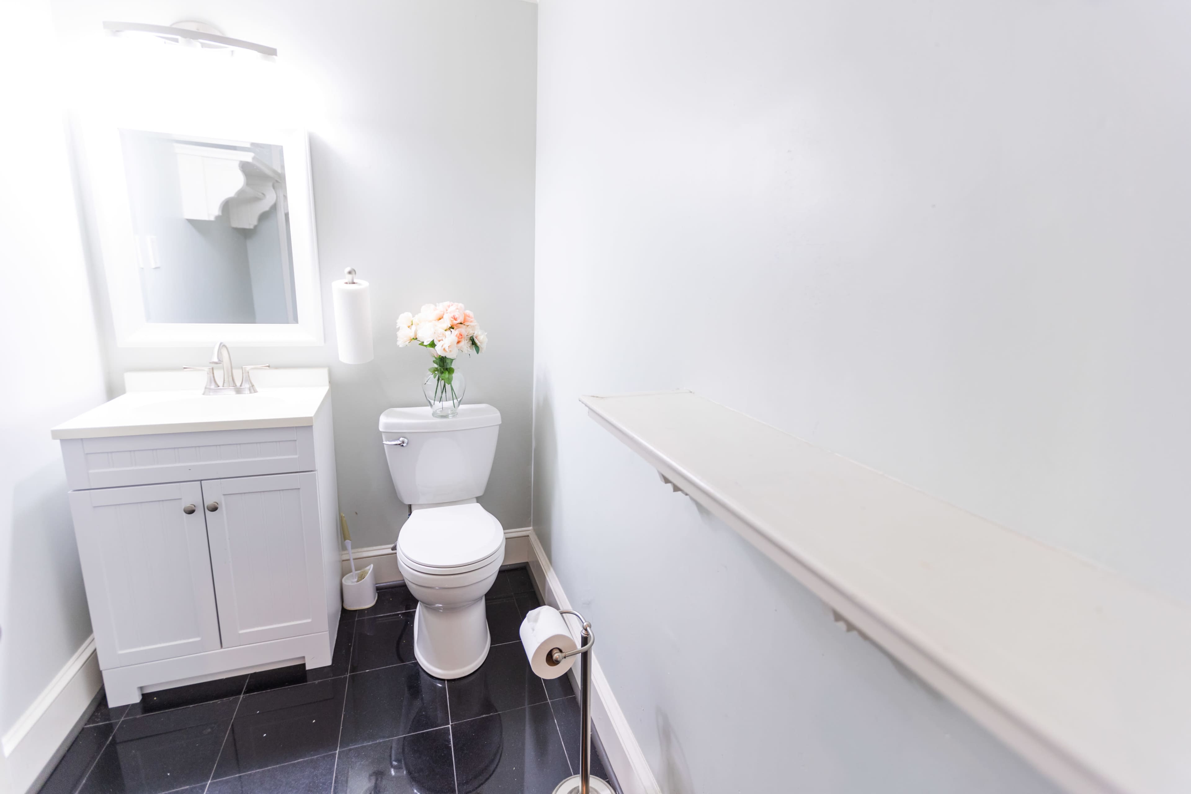 A clean bathroom with a white toilet, a small vanity with a sink, and a shelf along the wall, adorned with a flower arrangement.