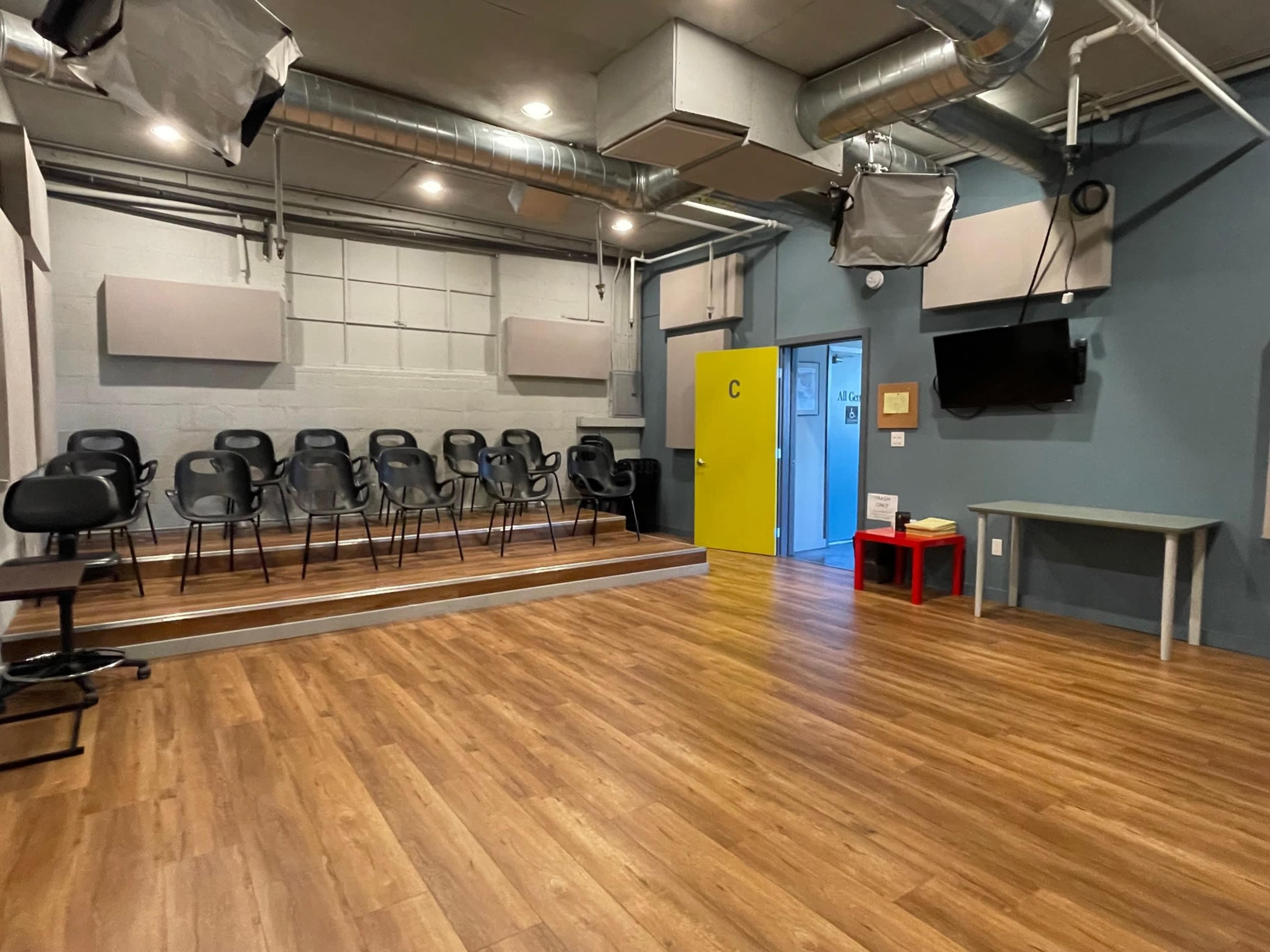 A spacious room with a wooden floor, featuring several black chairs arranged on a stage, a yellow door, and a table against the wall.