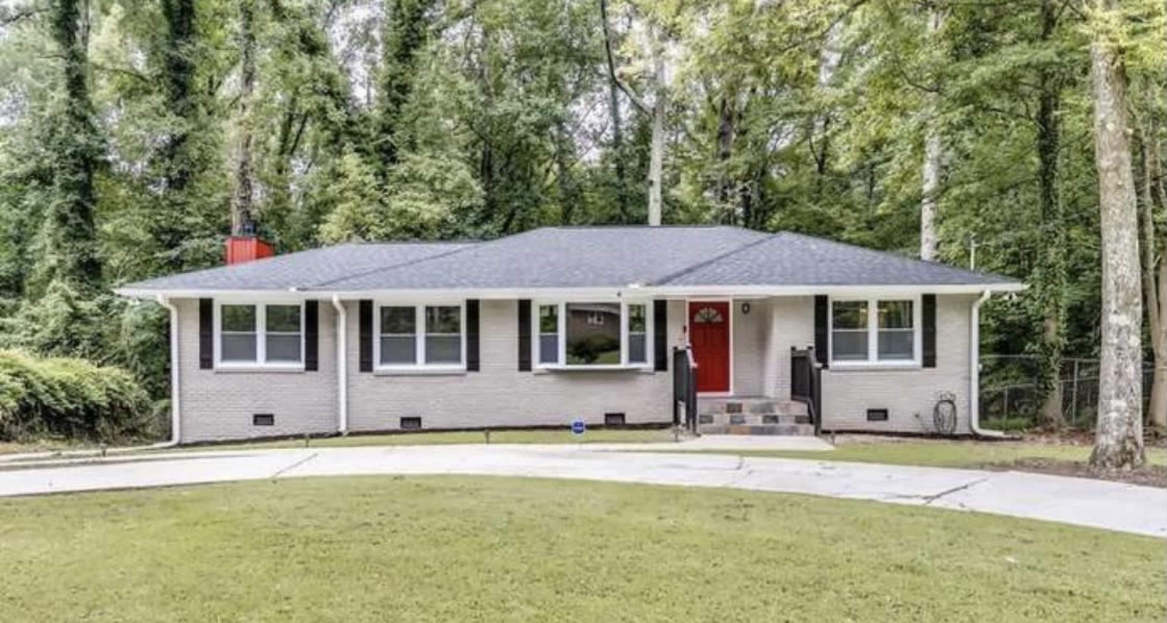 A single-story, modern home with a red front door, surrounded by greenery and a circular driveway.