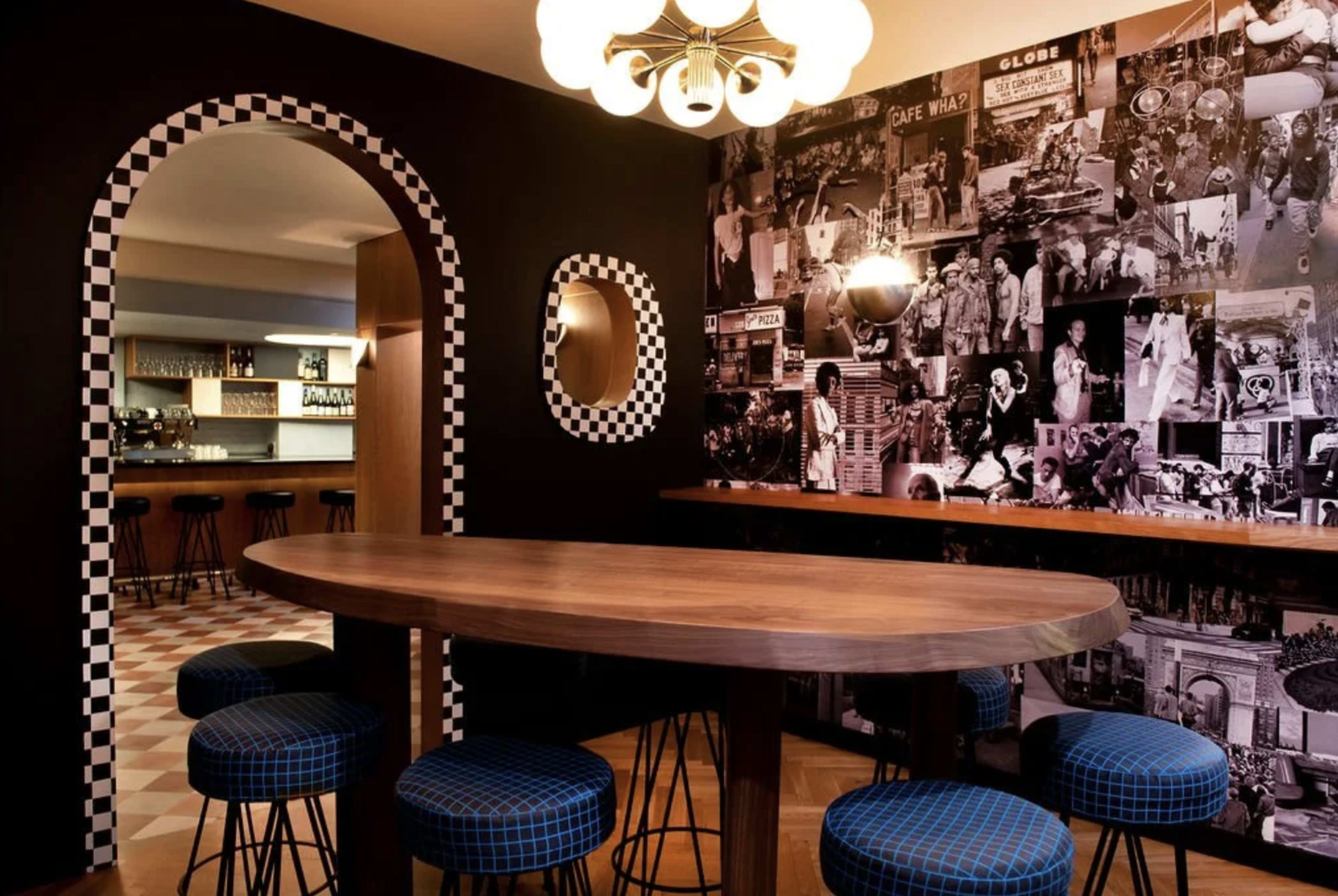 The image shows a modern dining area with a round wooden table surrounded by blue checkered stools, featuring a black and white photo collage wall and an archway.