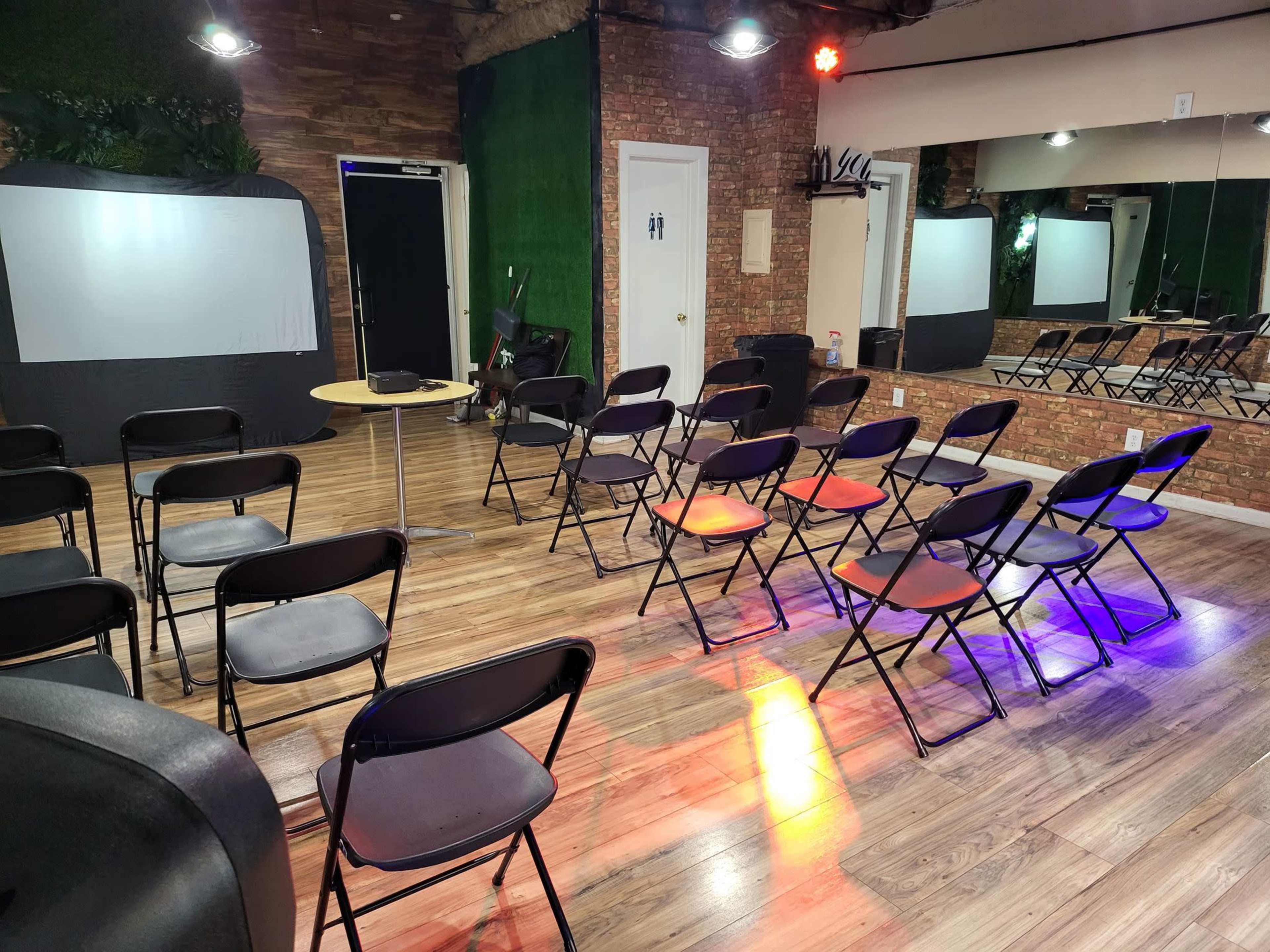 The image shows a room set up with several black folding chairs arranged in rows, a small round table, and a projection screen against a brick wall, with mirrors and colorful lighting in the background.