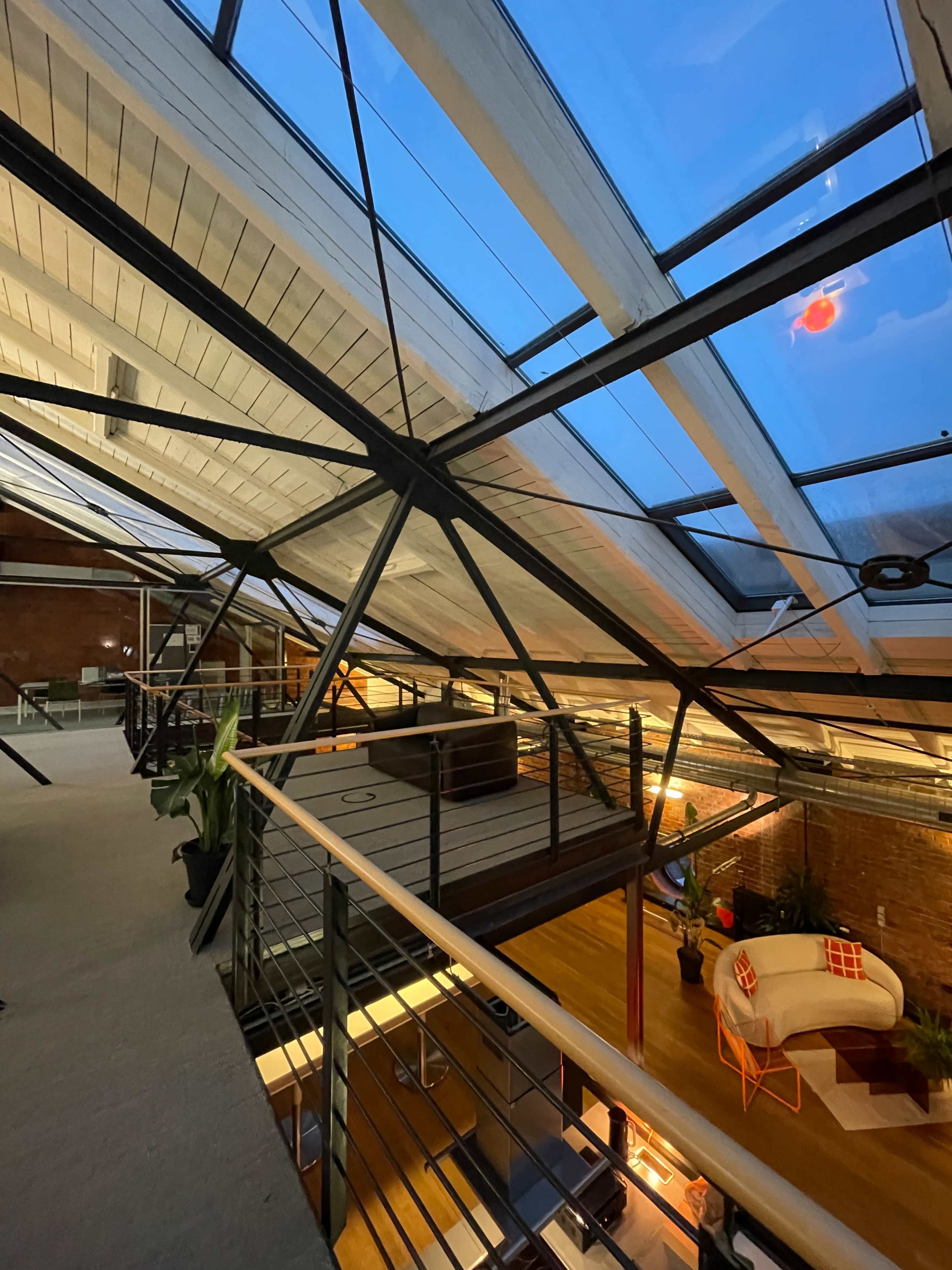 The image shows an industrial-style loft with a high ceiling, visible steel beams, and large skylights allowing natural light to illuminate the space.