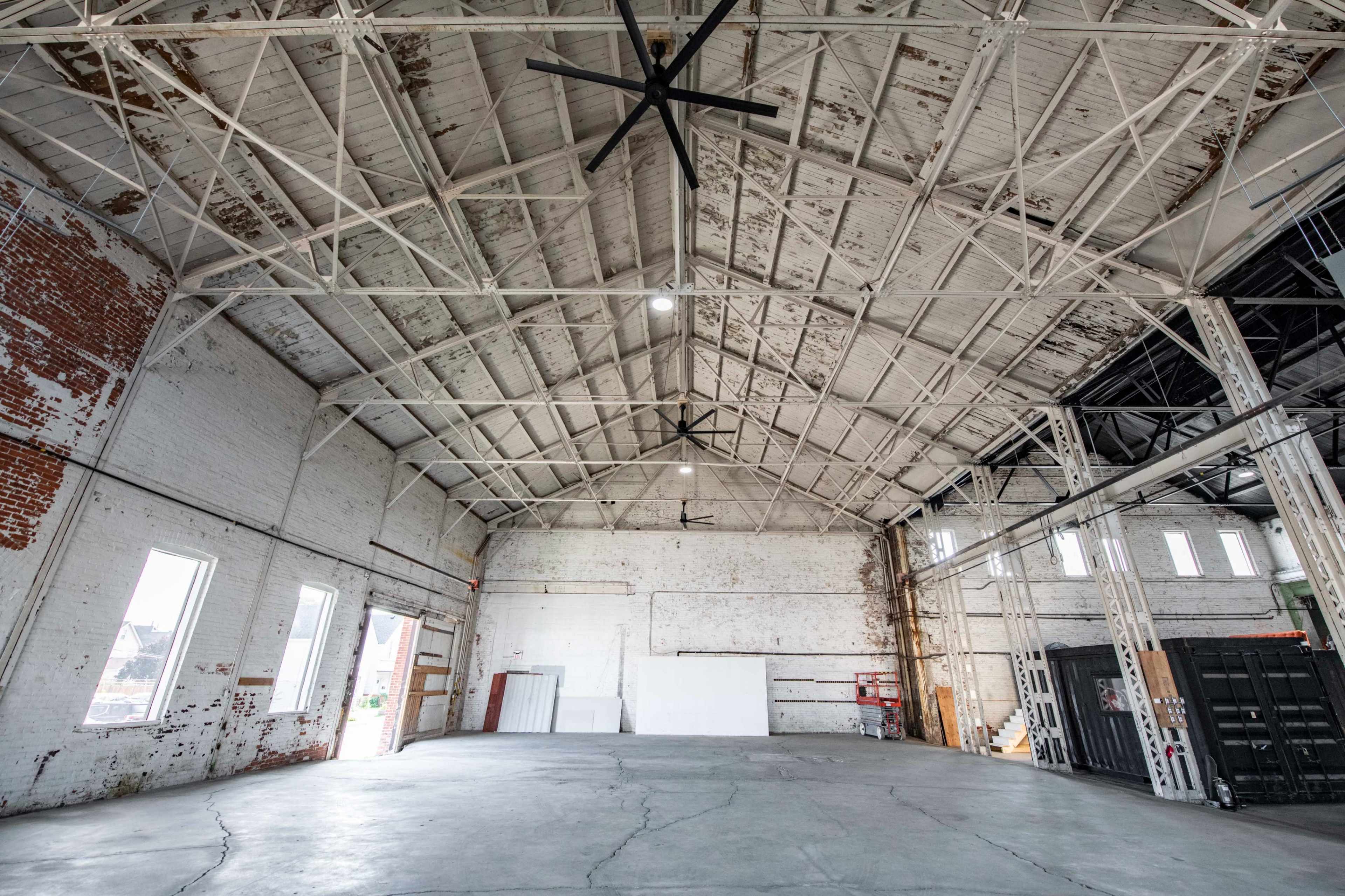 The image shows an empty industrial warehouse with a high ceiling, exposed brick walls, and large windows.