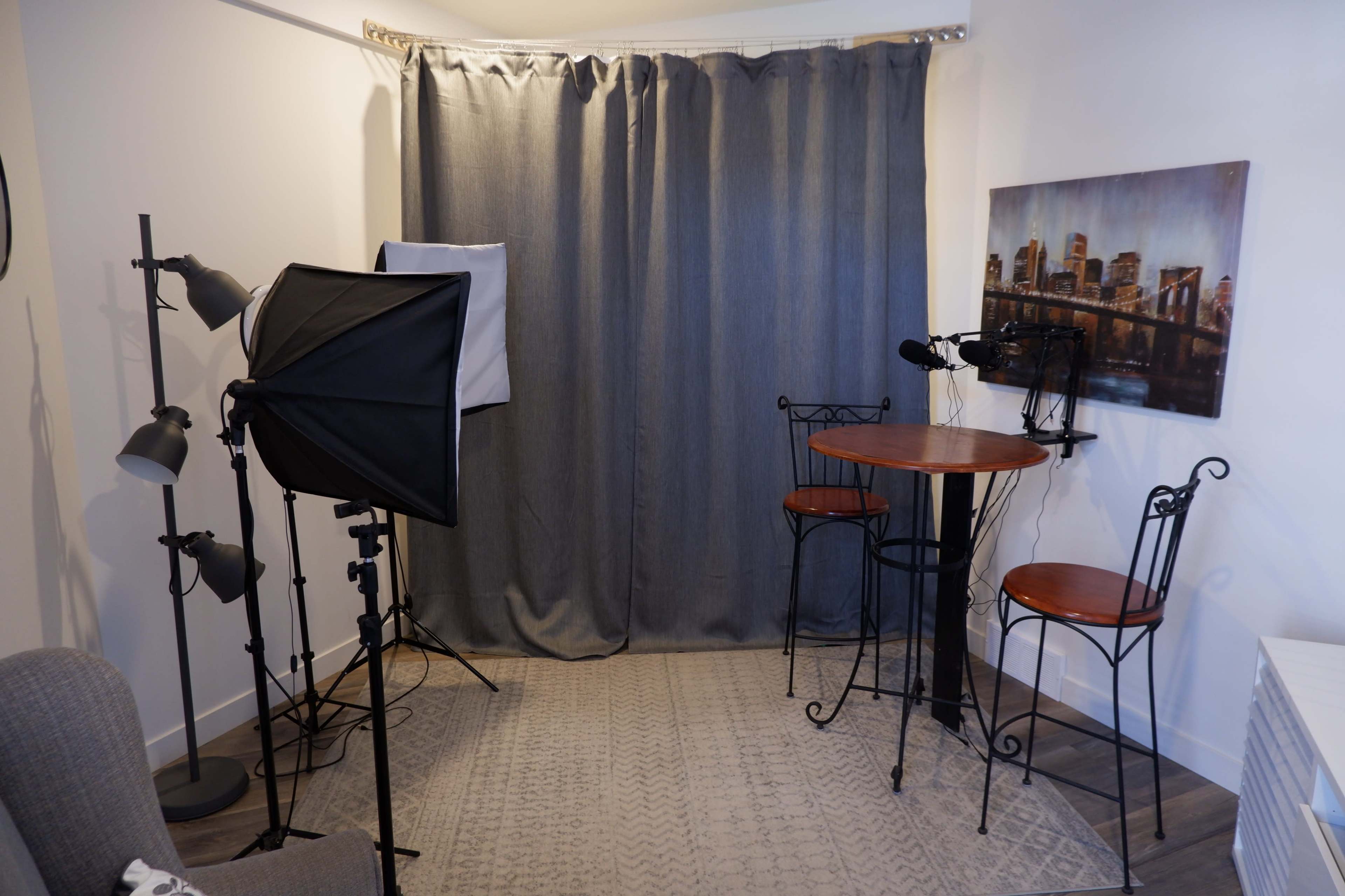 The image shows a small studio setup featuring a gray curtain backdrop, two bar stools at a tall table, and various lighting equipment arranged nearby.