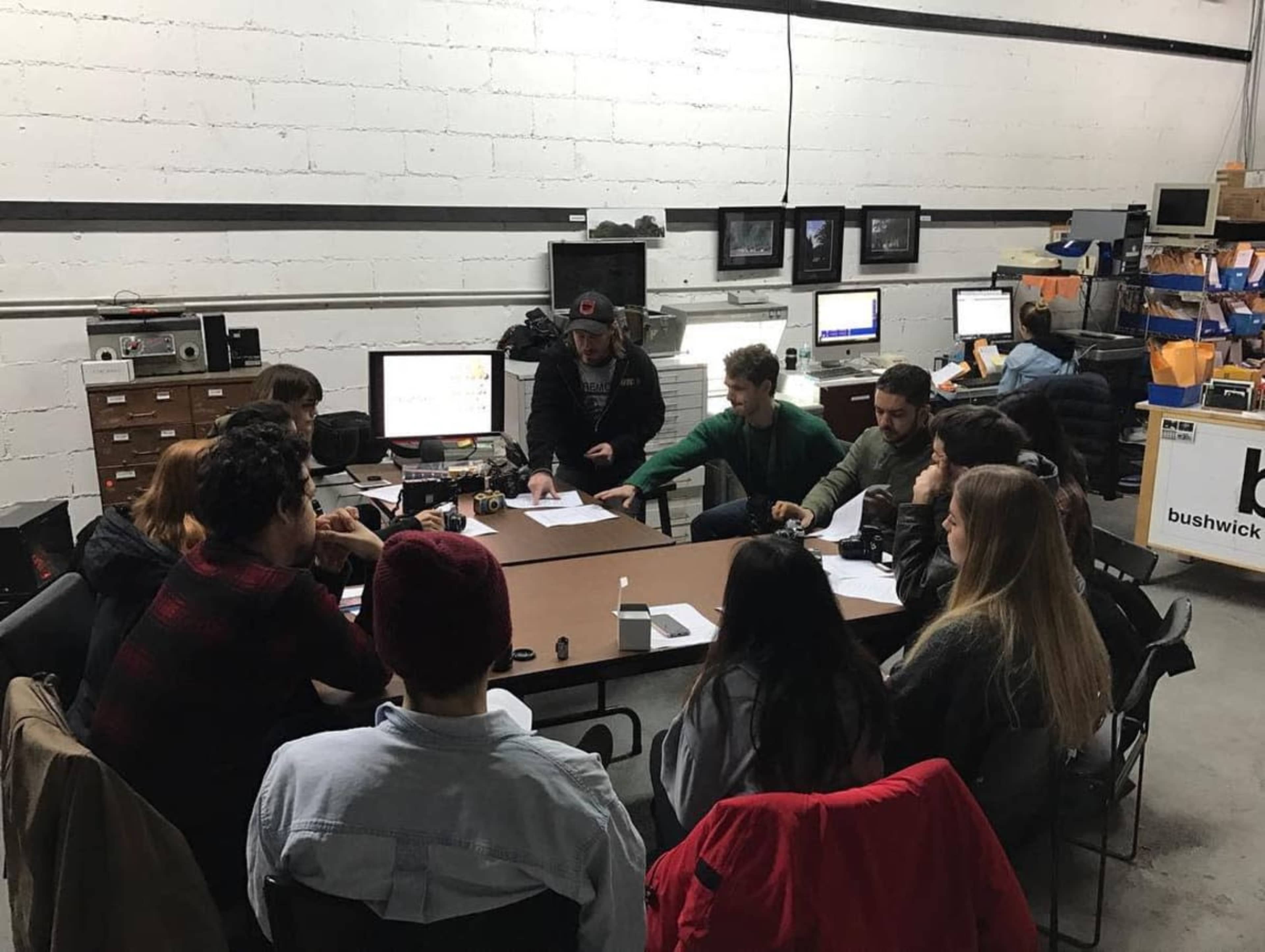 A group of twelve people is gathered around a table in a warehouse, with computers and equipment visible in the background.