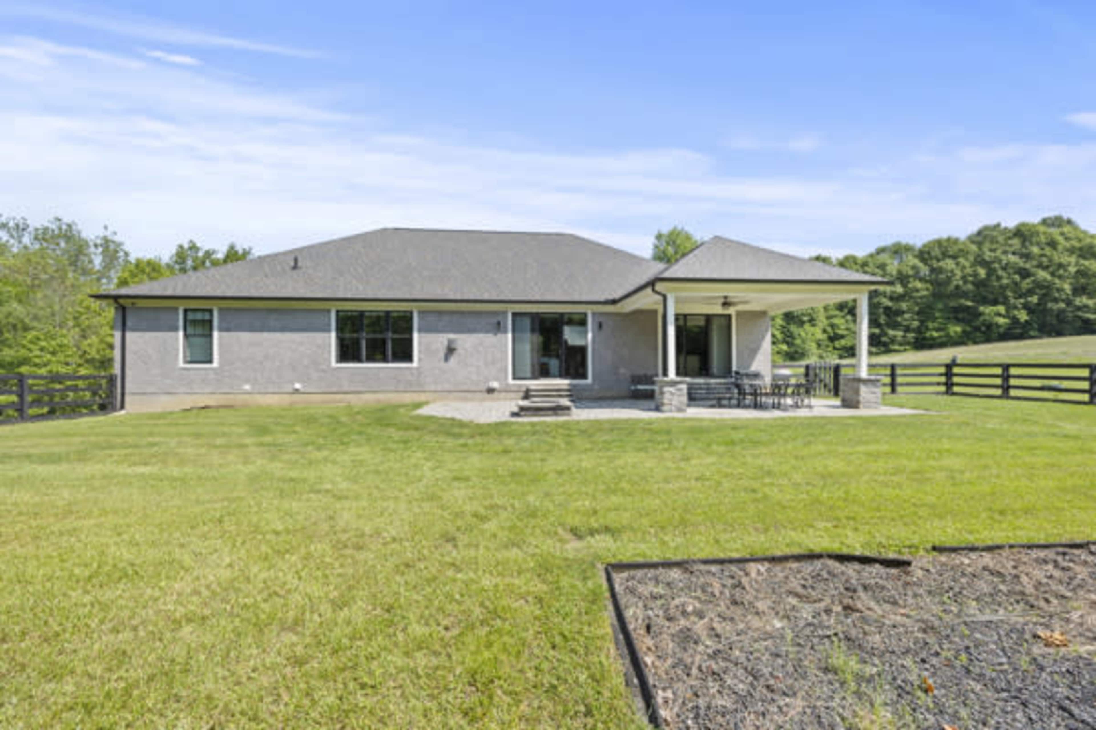 The image shows a modern single-story house with a covered patio and a large lawn, surrounded by trees and open fields.