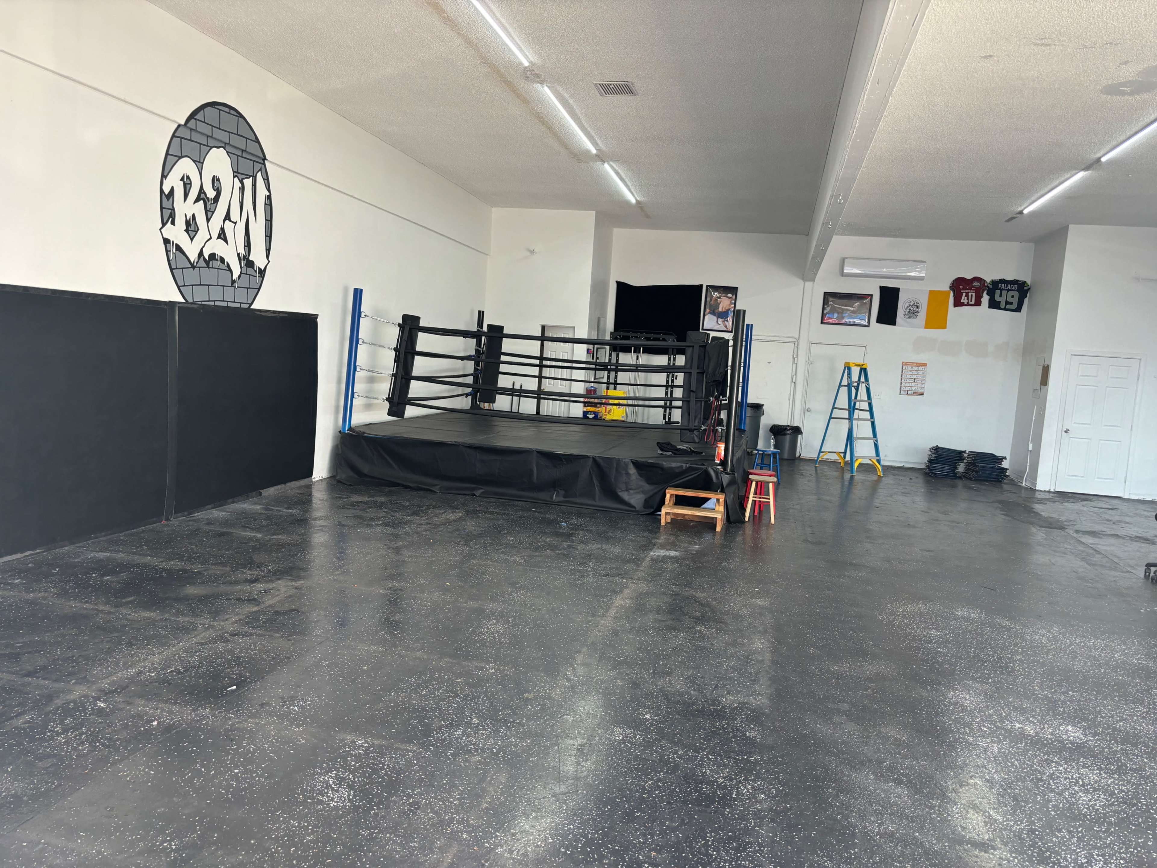 The image shows an empty boxing gym featuring a boxing ring, black walls, and various pieces of equipment like a ladder and chairs.