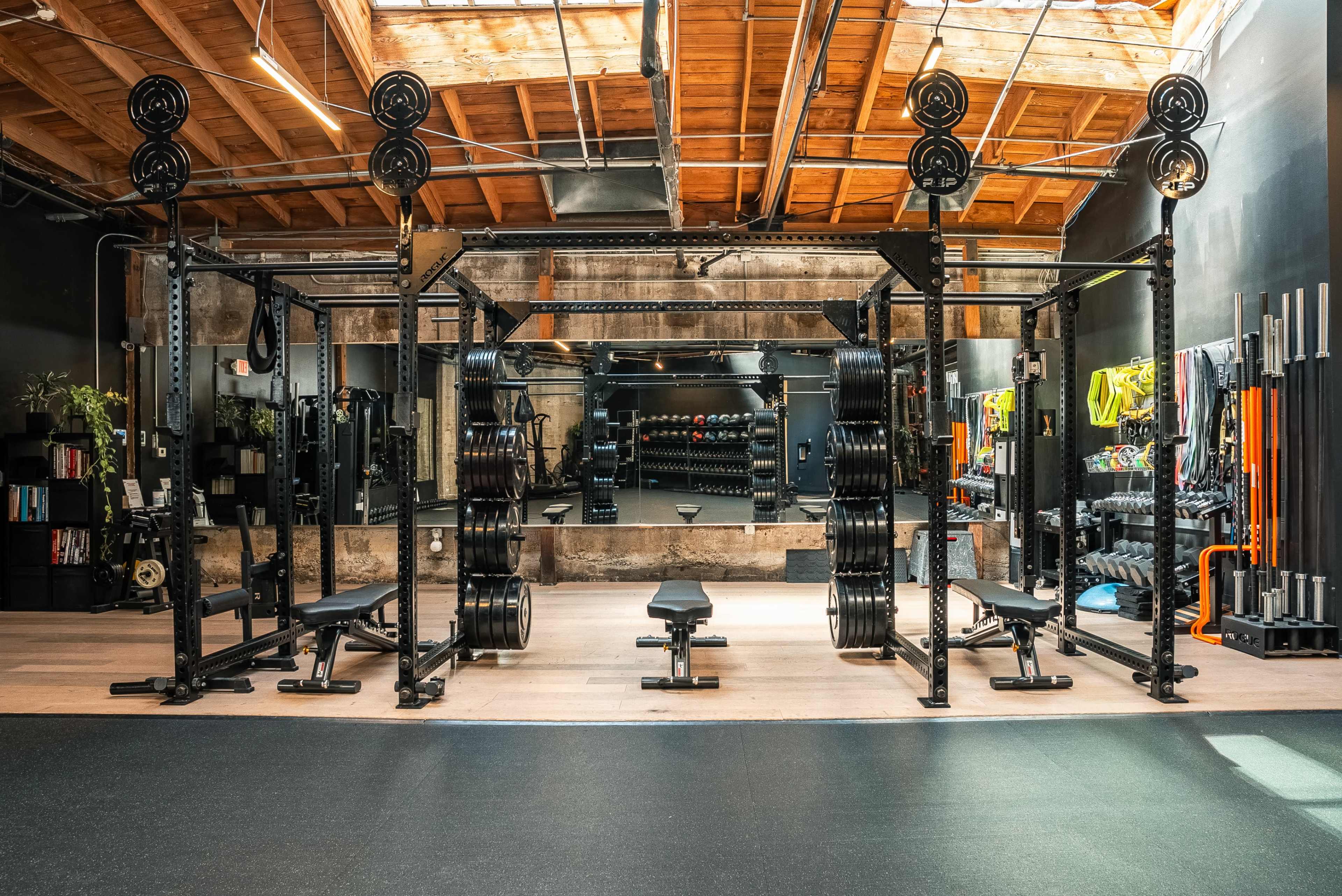 The image shows a fitness facility featuring a weightlifting rack with weights, a bench press, and various exercise equipment, all arranged in a well-lit space with a mirrored wall.