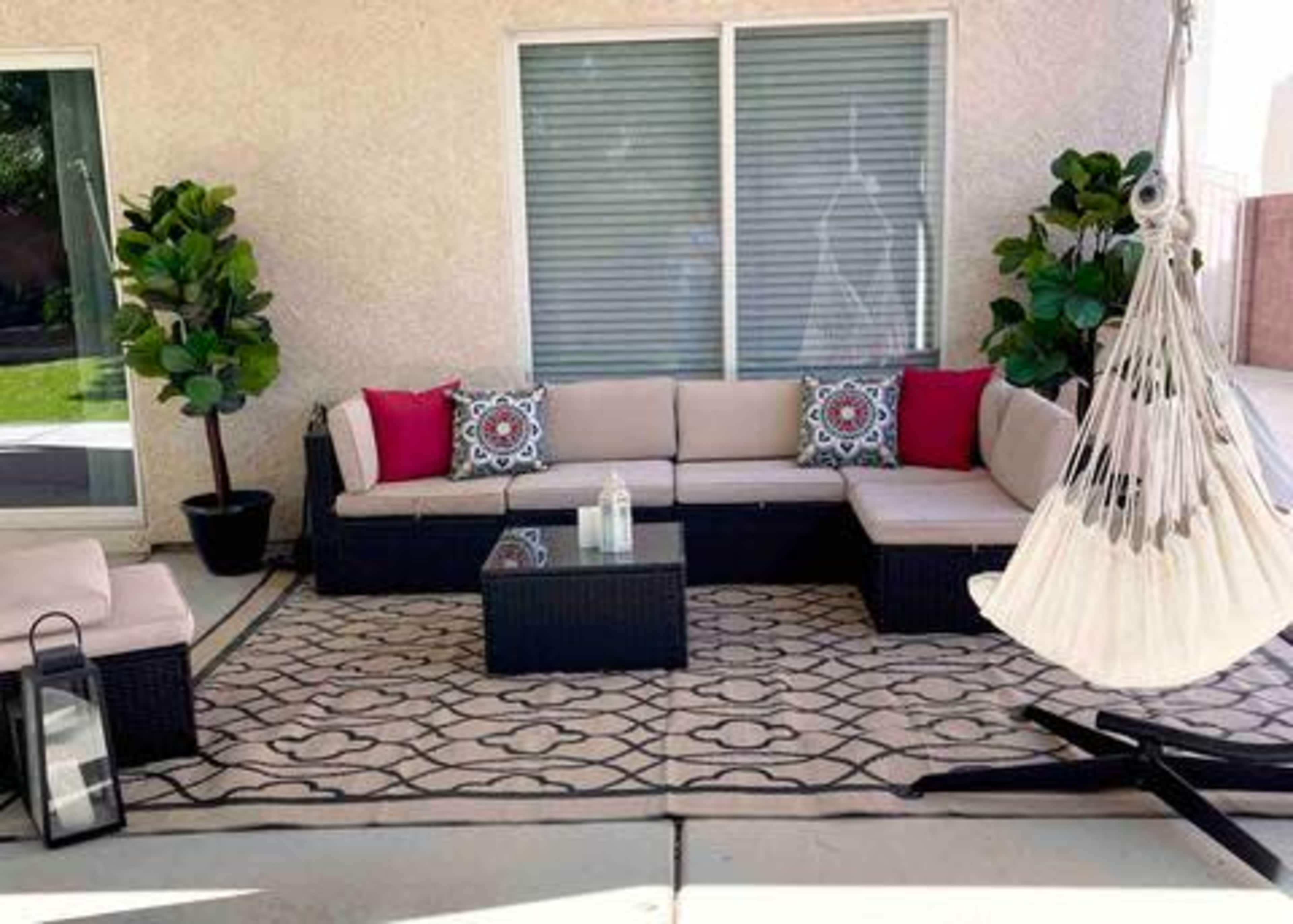 The image shows a patio area with a sectional couch, decorative pillows, a glass coffee table, a hammock, and potted plants.