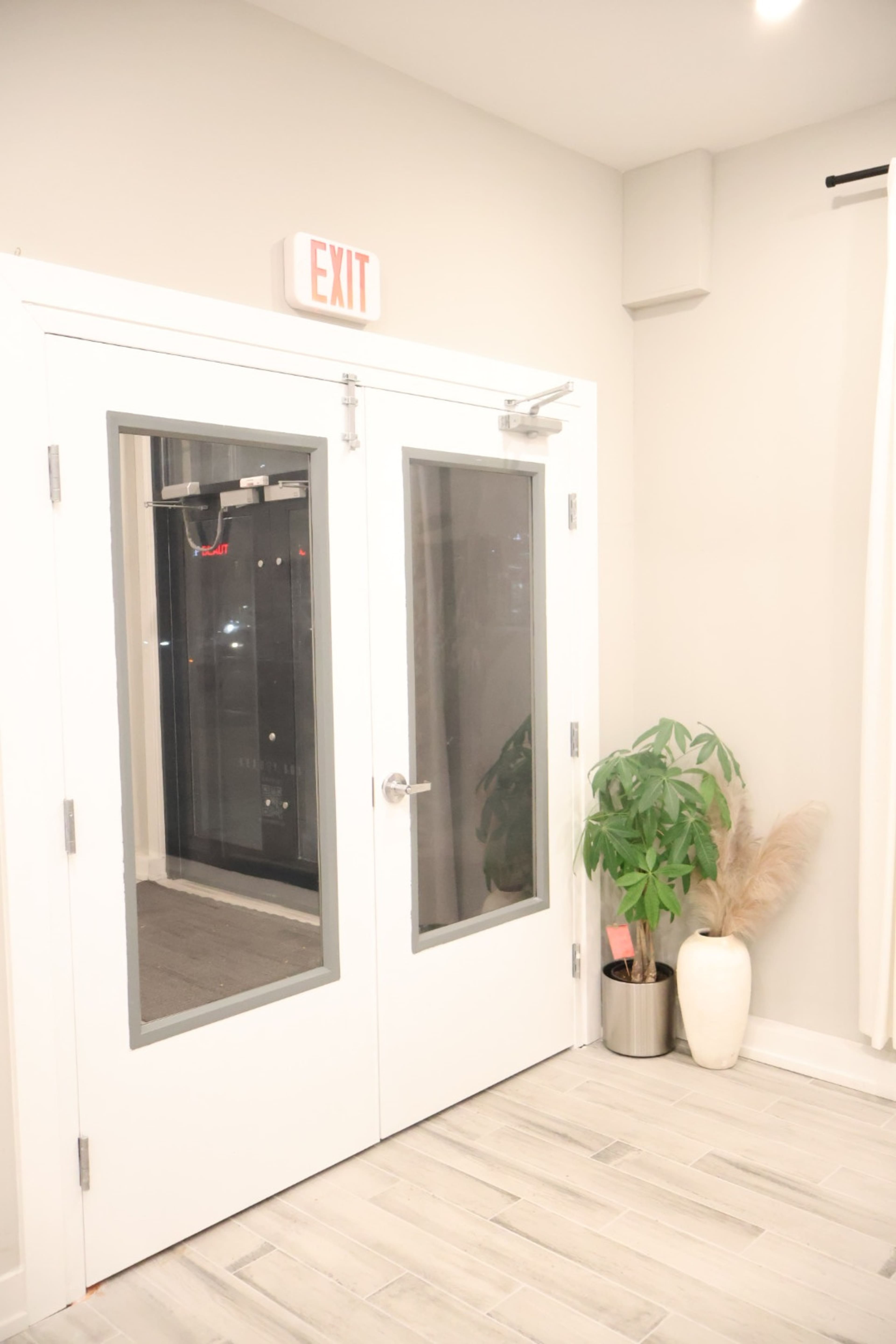 The image shows a pair of glass double doors marked with an "EXIT" sign, adjacent to a green potted plant and a large white vase, in a well-lit room with light-colored walls and tiled flooring.
