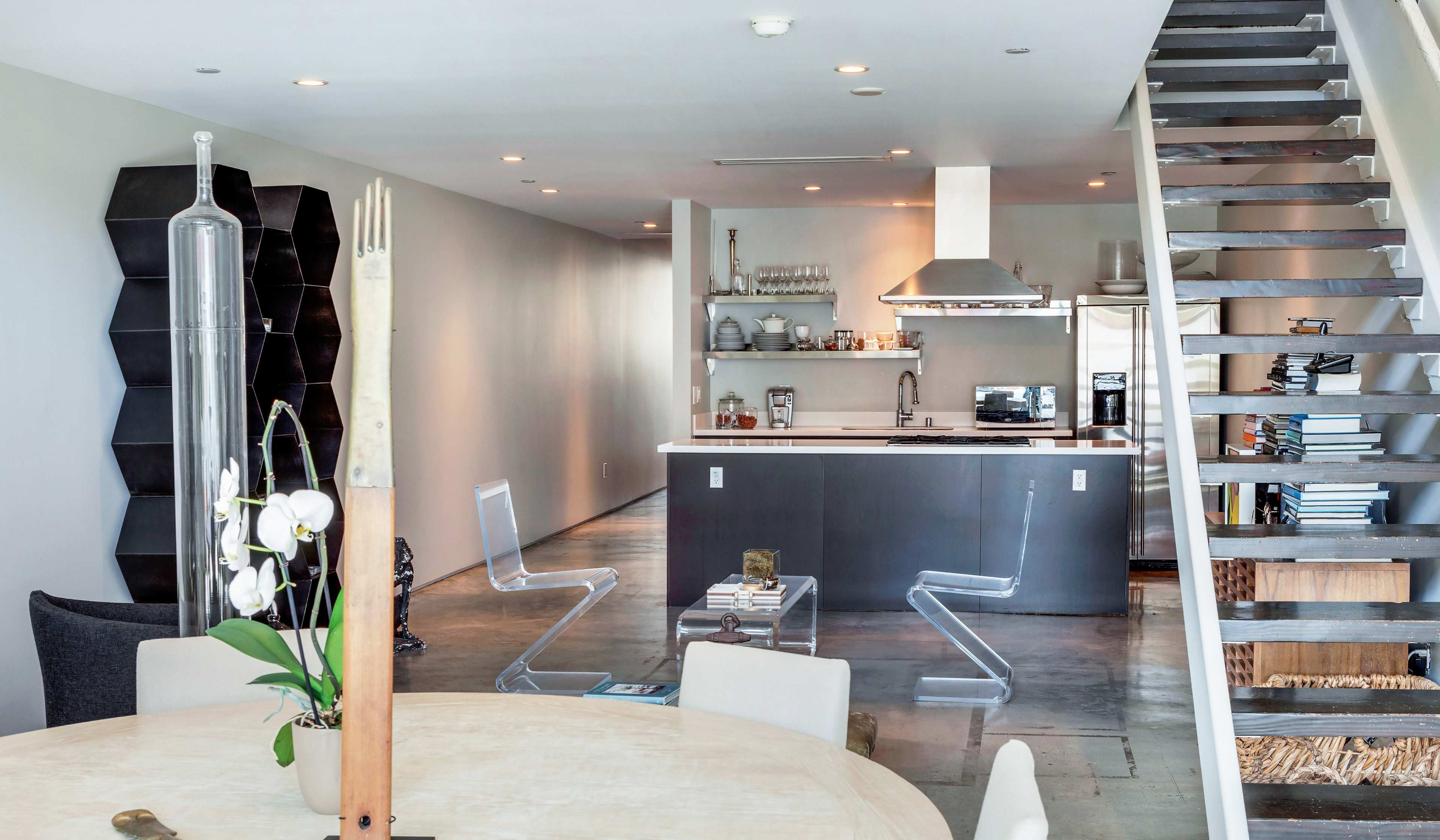 The image shows a modern open-concept living space featuring a kitchen with a central island, a dining table, transparent chairs, and a staircase leading to an upper level.