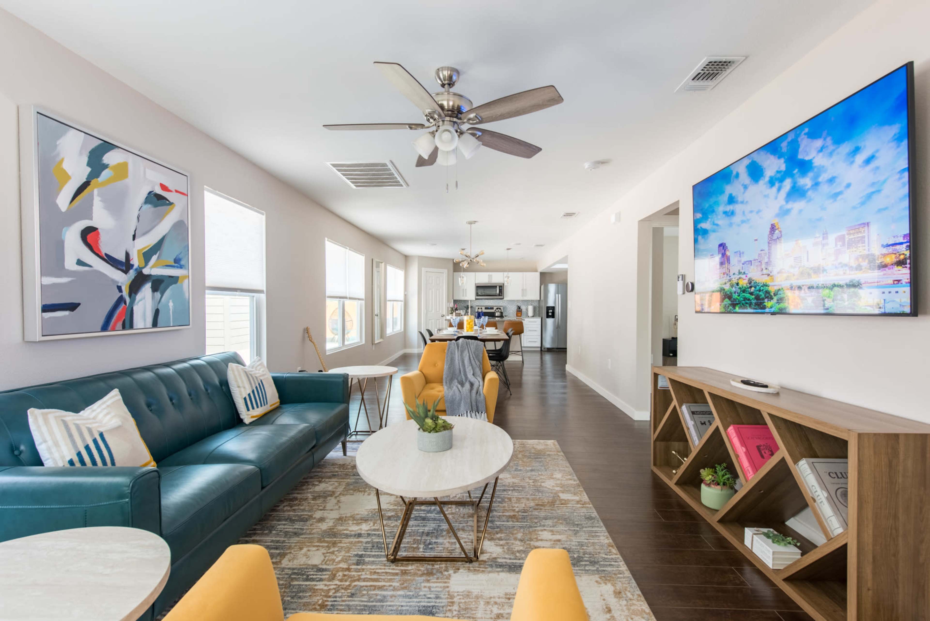 The image shows a modern living room with a teal sofa, a marble coffee table, and a large wall-mounted TV, adjacent to a kitchen visible in the background.
