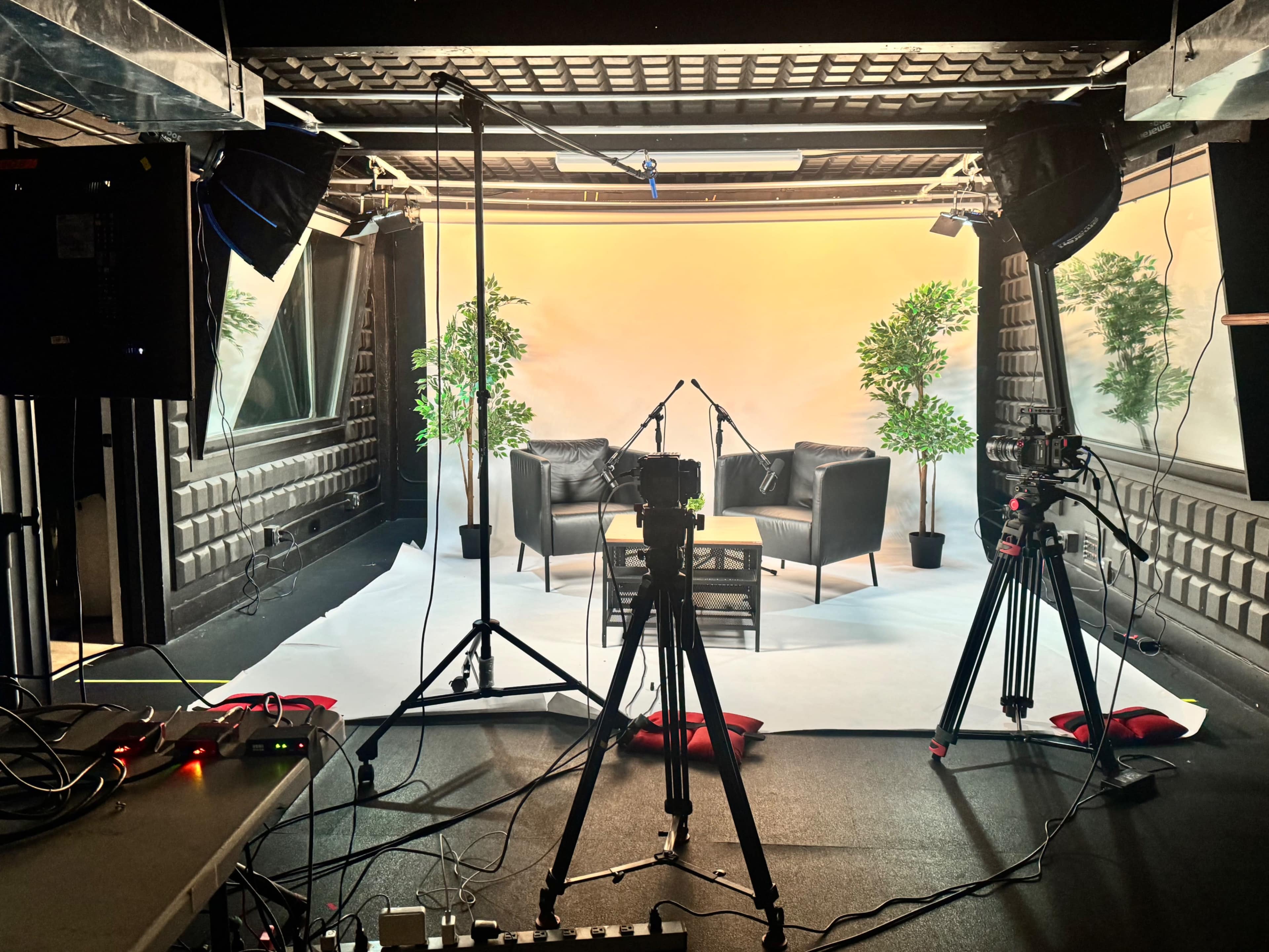 The image shows a film studio setup with two black chairs, a coffee table, and potted plants arranged against a backdrop.