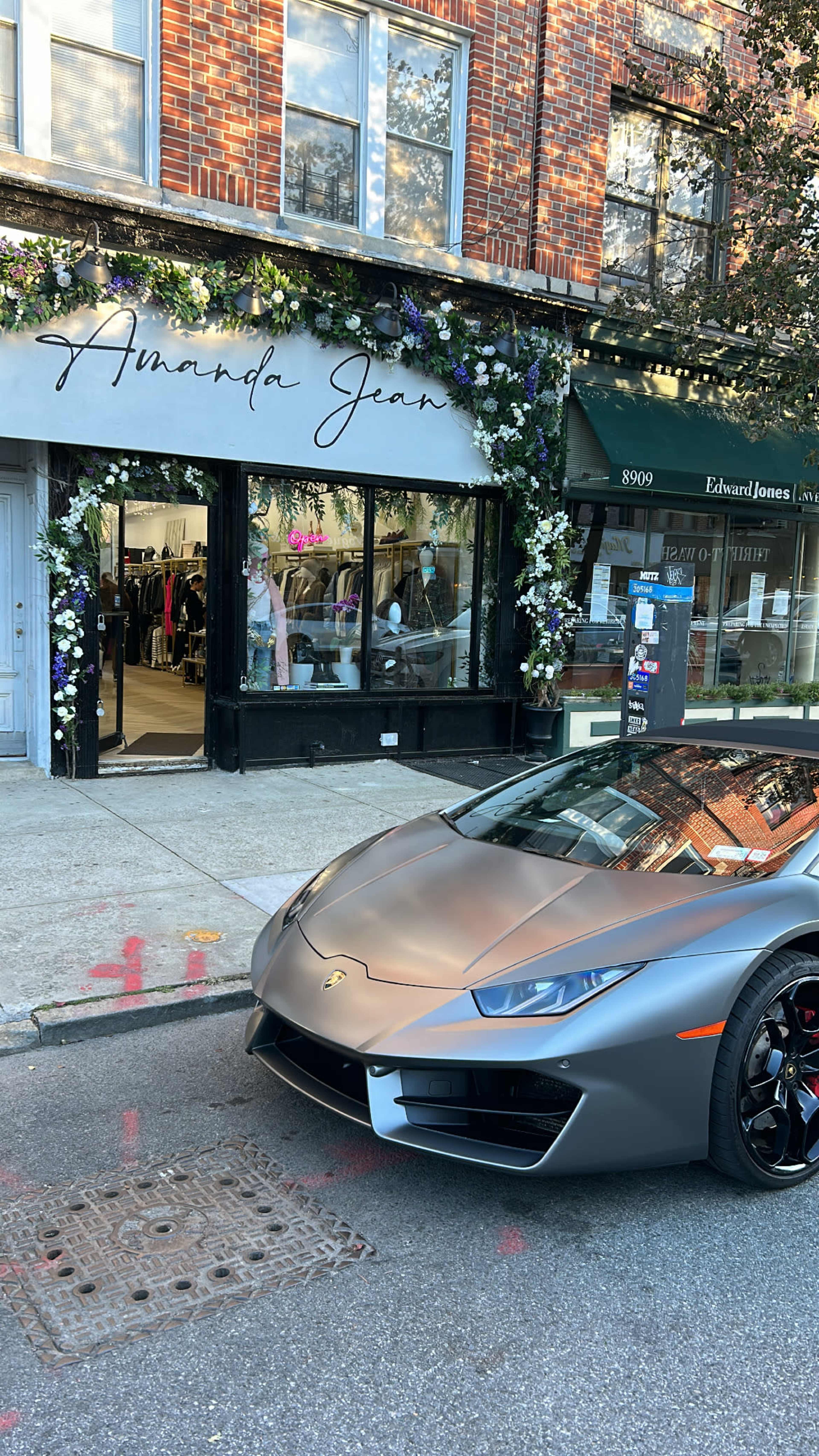 A sleek gray sports car is parked in front of a boutique named "Amanda Jean," which has floral decorations on its exterior.