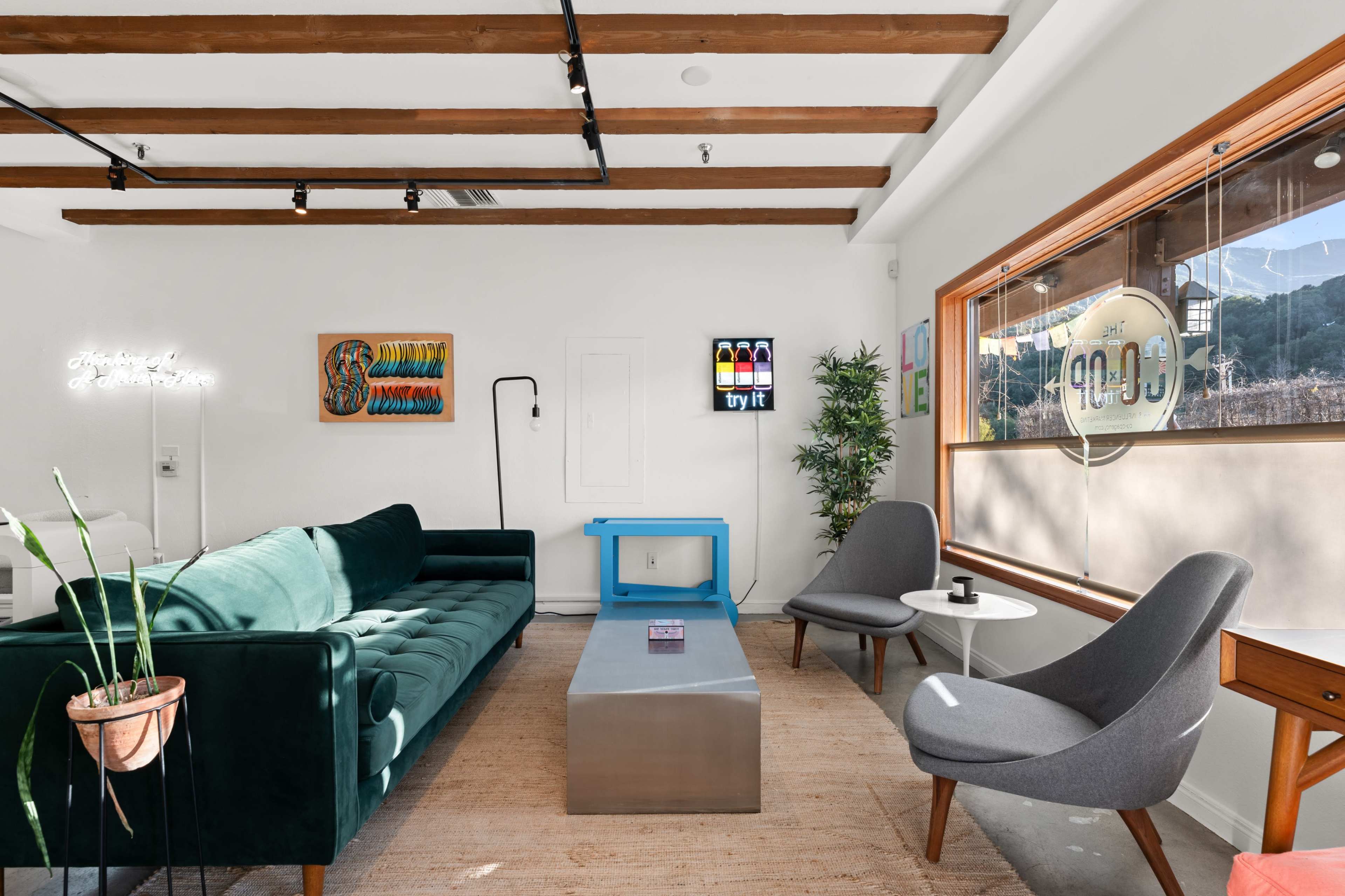 A modern, minimalist room featuring a green sofa, a metal coffee table, and two gray chairs, with natural light coming through a large window.