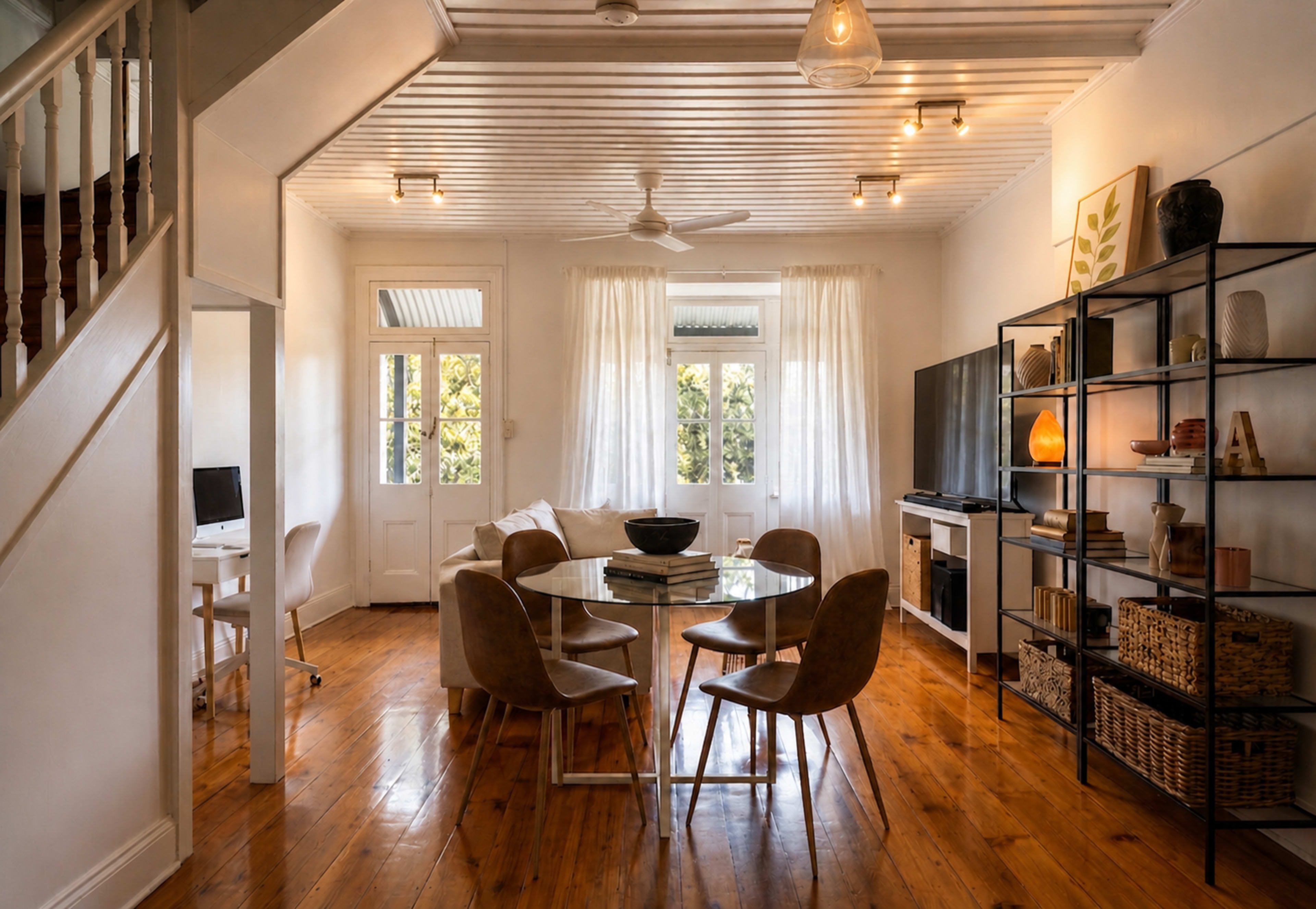 The image shows a modern living space with a glass dining table surrounded by brown chairs, a television on a black shelving unit, and large windows with white curtains.