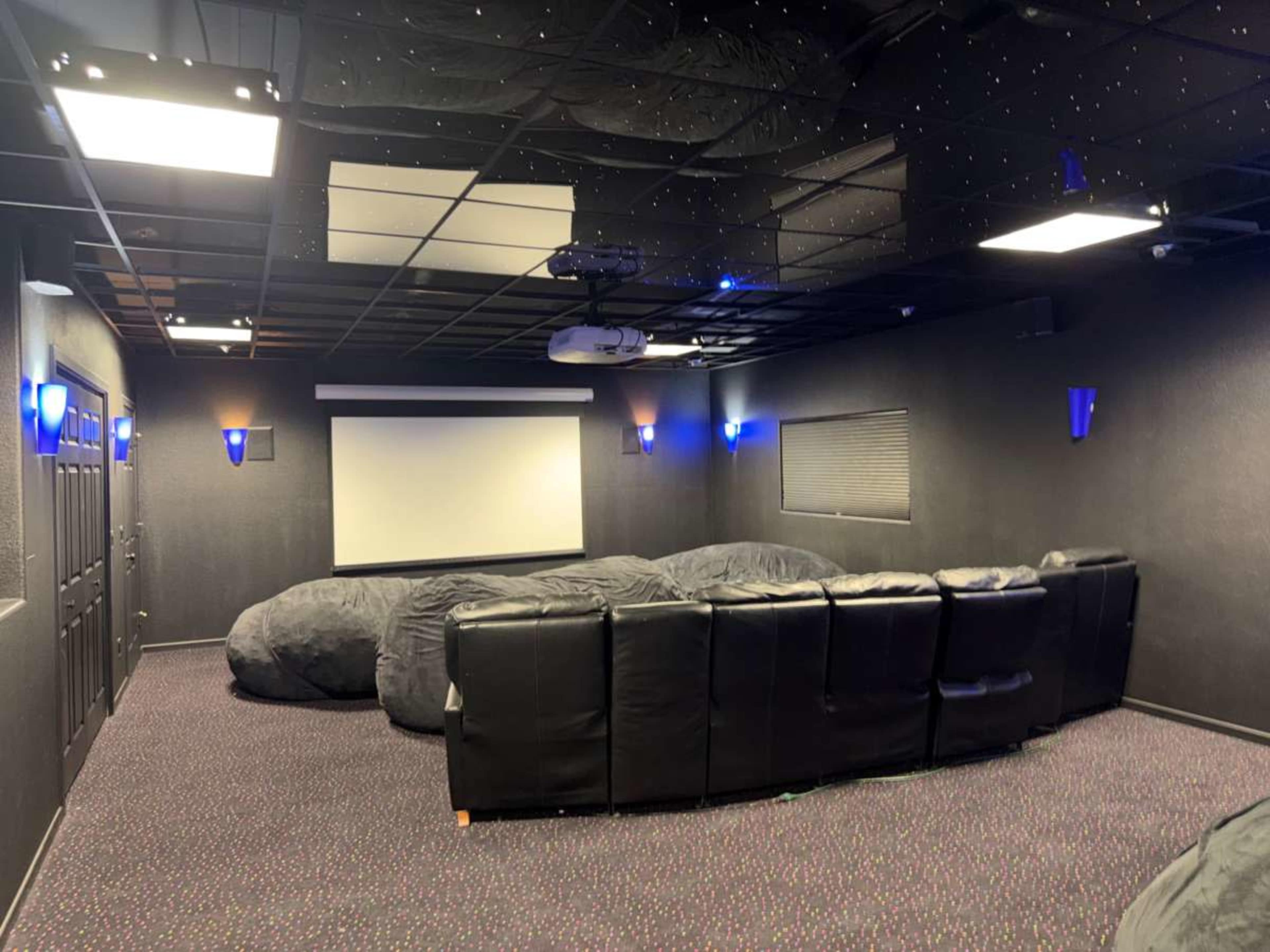 A home theater room features black walls, a large screen at the front, a couch along the back, and several large bean bag chairs scattered on the floor.
