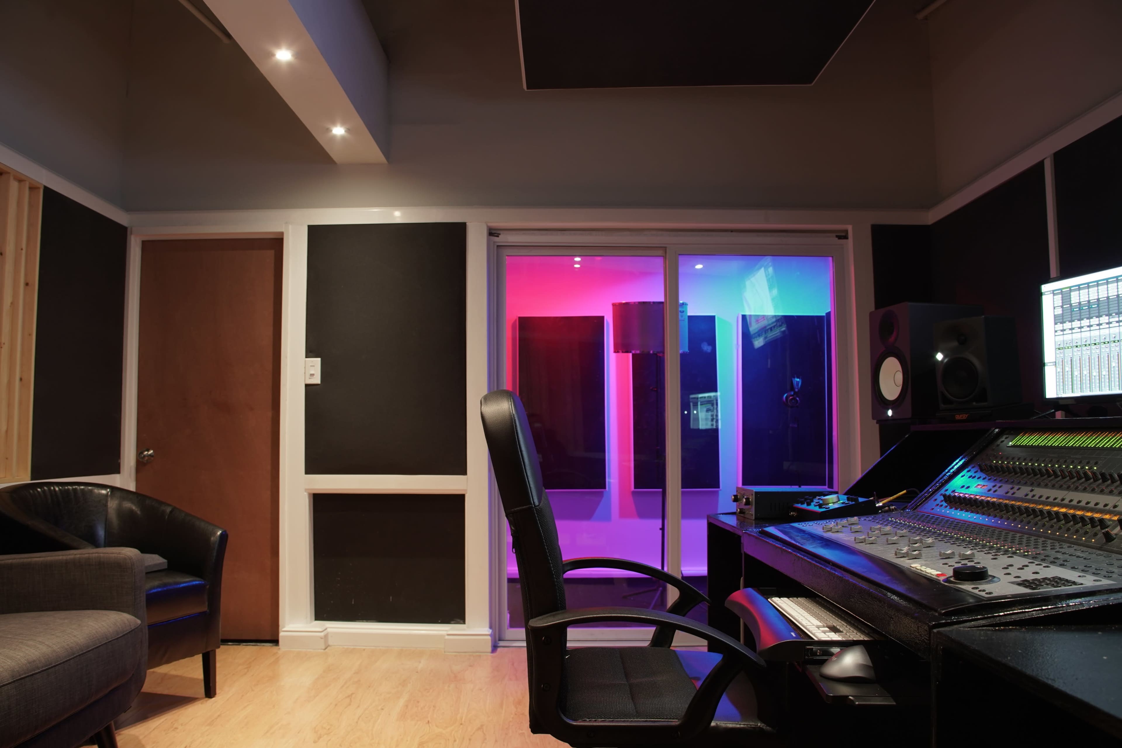 A recording studio with a mixing console, a chair, and a glass-enclosed sound booth illuminated in blue and pink lighting.