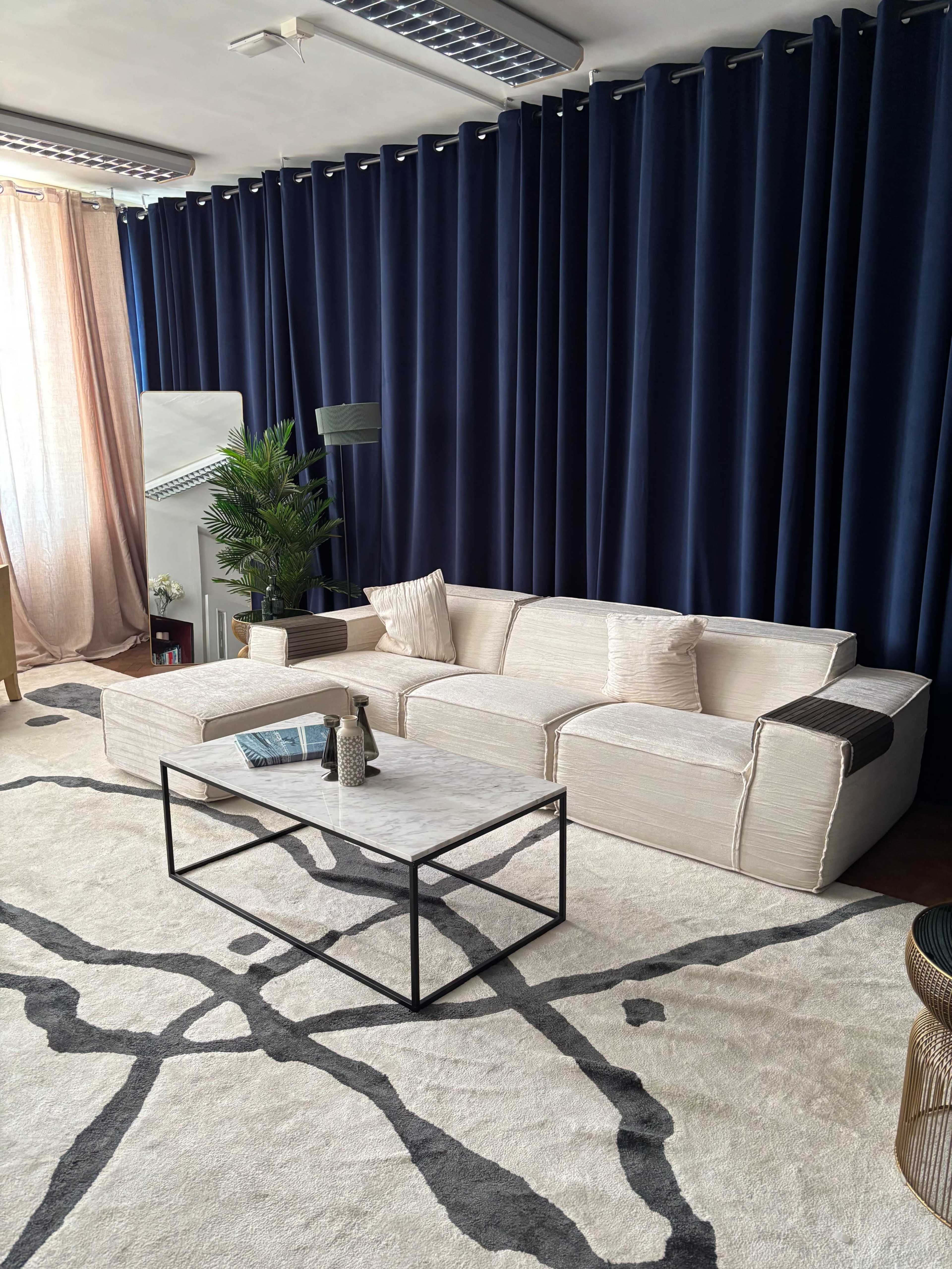 The image shows a modern living room with a light-colored sectional sofa, a marble-top coffee table, and large dark curtains draping a window.