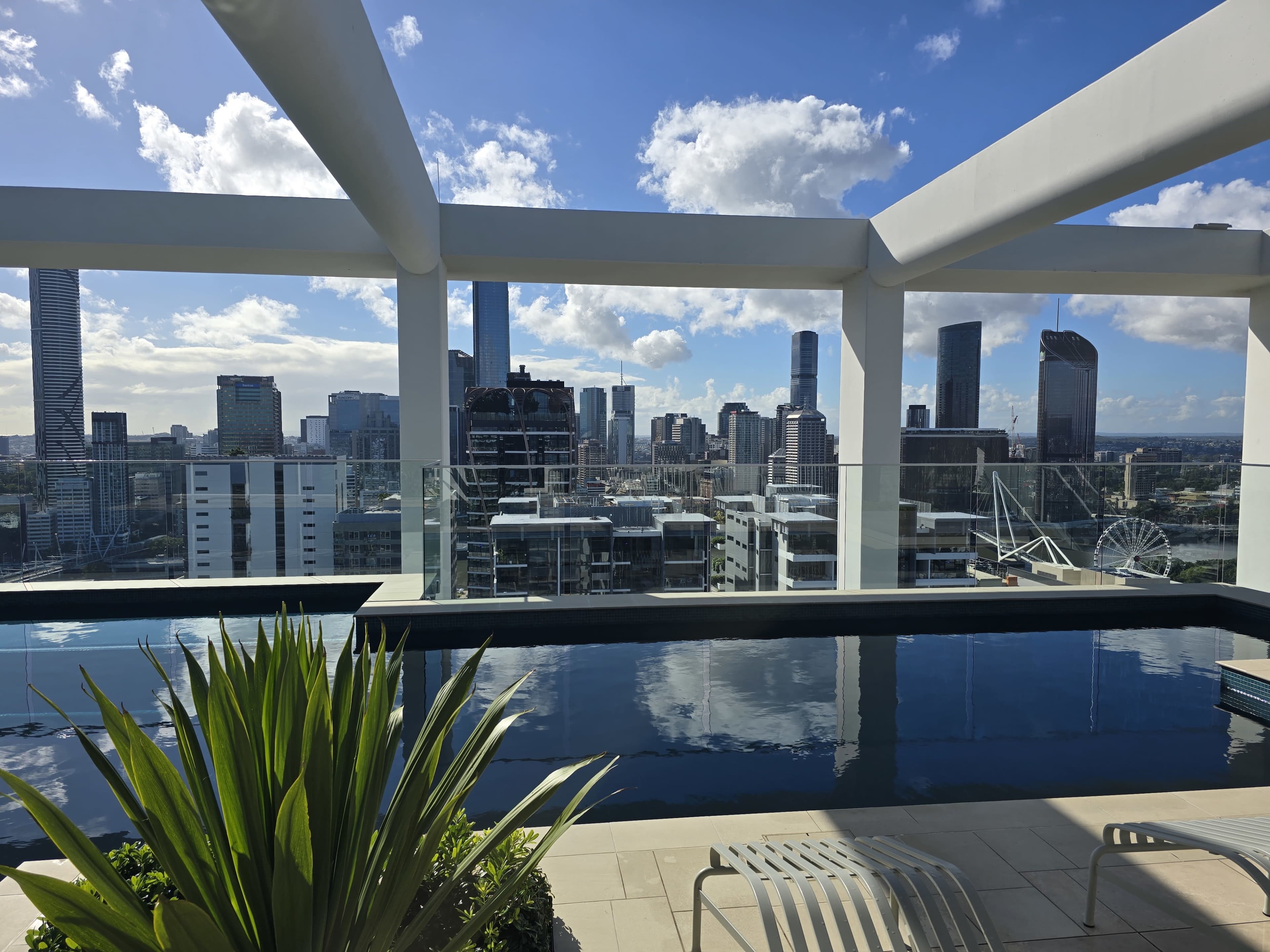 The image shows a rooftop pool surrounded by modern architecture, with a city skyline and cloudy blue sky in the background.