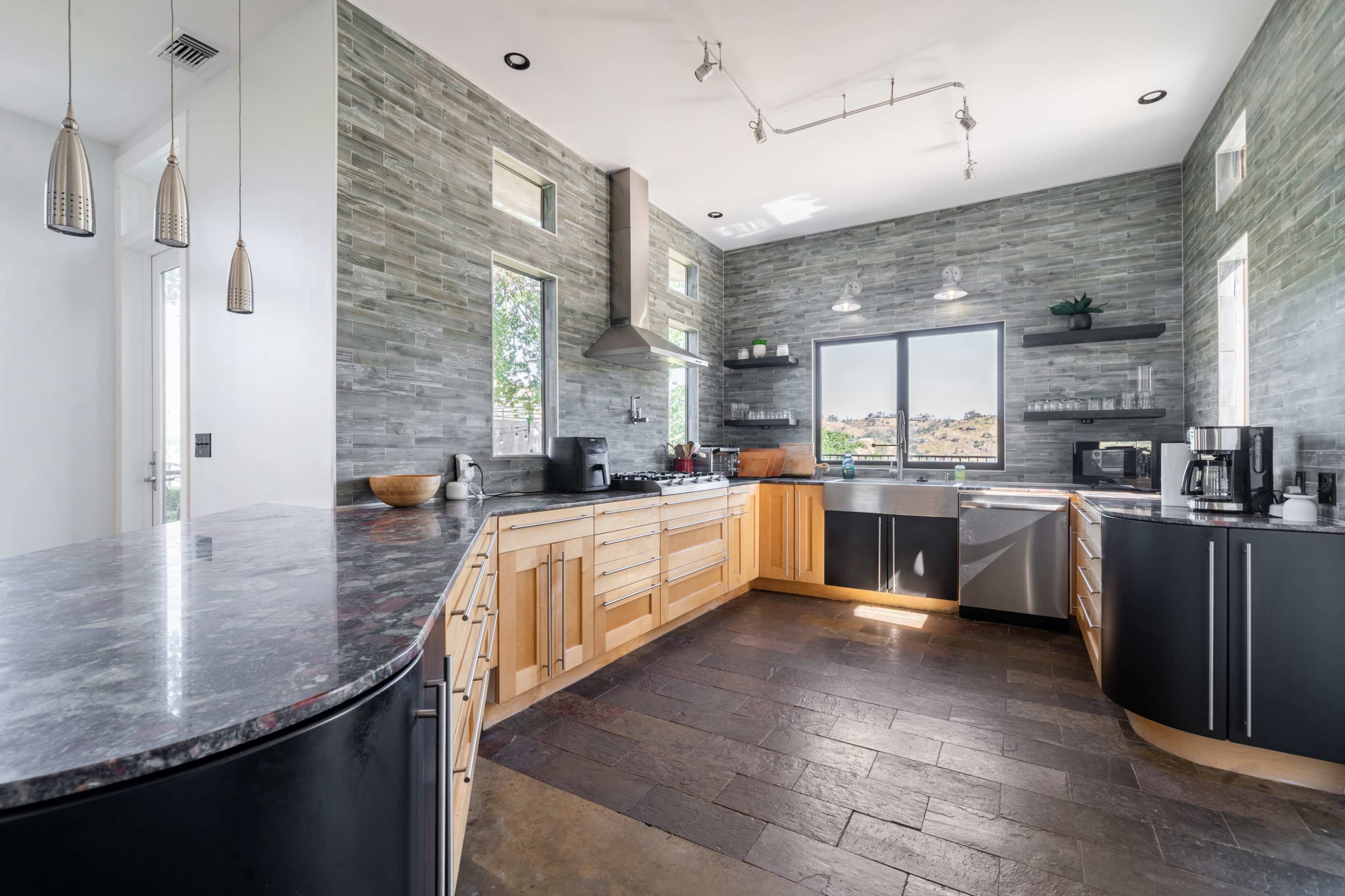 The image shows a modern kitchen featuring a combination of wooden and metallic cabinetry, large windows providing natural light, and a black stone countertop.