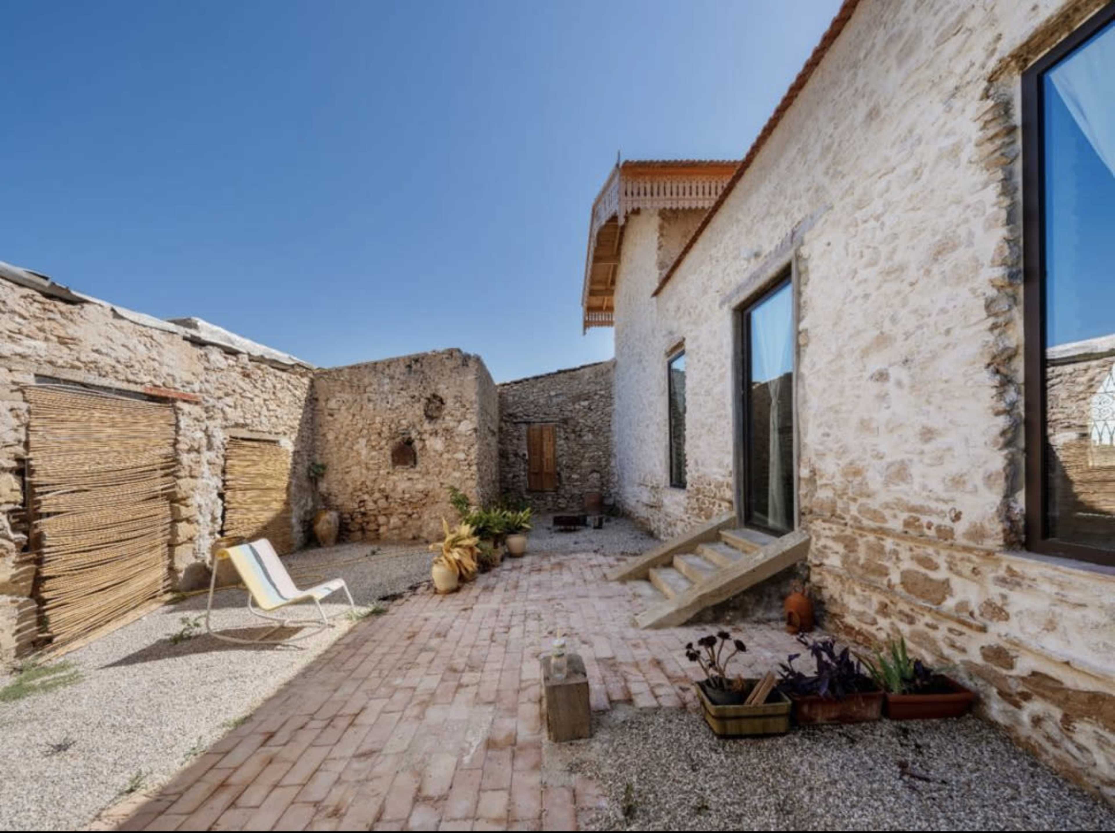 The image shows a stone courtyard with a pathway leading to a building featuring large windows and a wooden overhang, surrounded by various plants and a lounge chair.