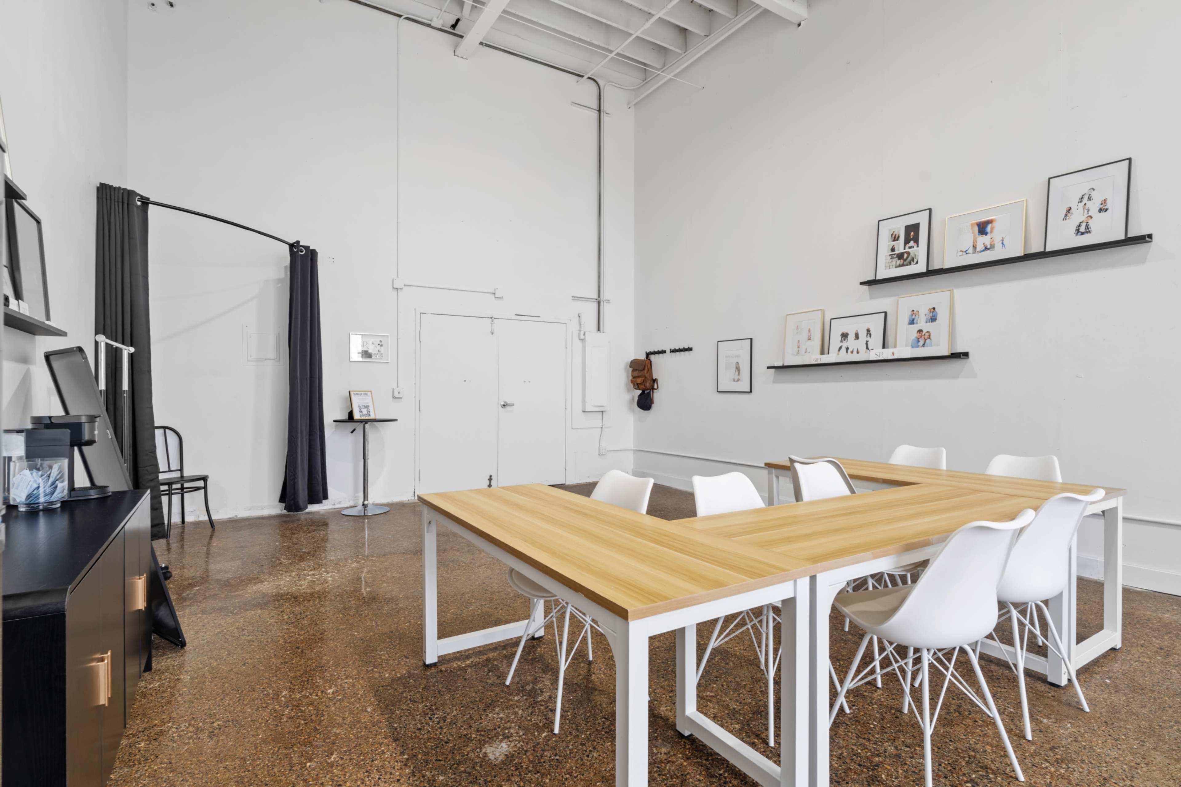 The image shows a spacious, minimalist meeting room with a large wooden table surrounded by white chairs, featuring a few framed artworks on the wall and a curtain to one side.