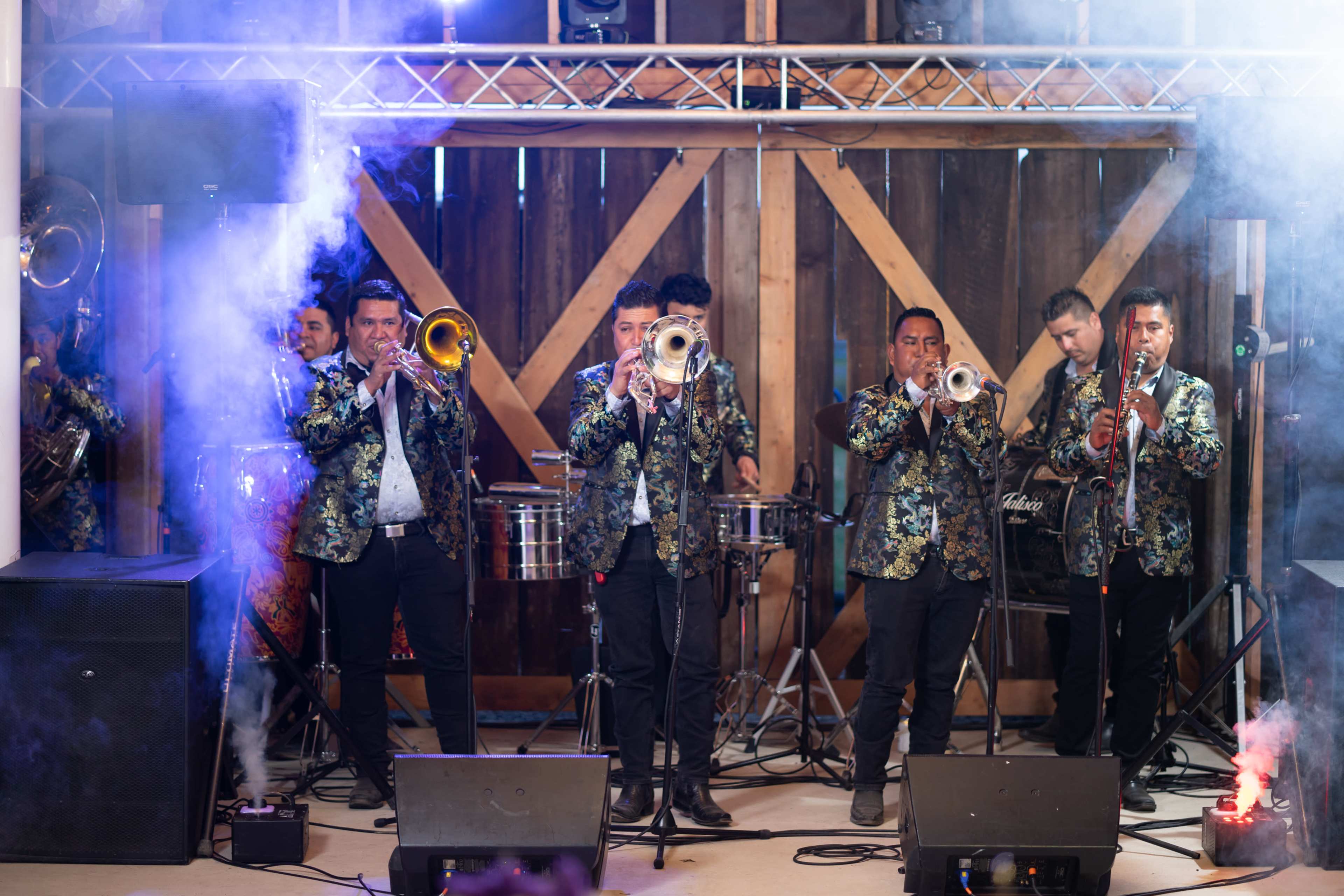 A band of musicians in ornate jackets plays brass instruments on a stage surrounded by smoke and colorful lighting.