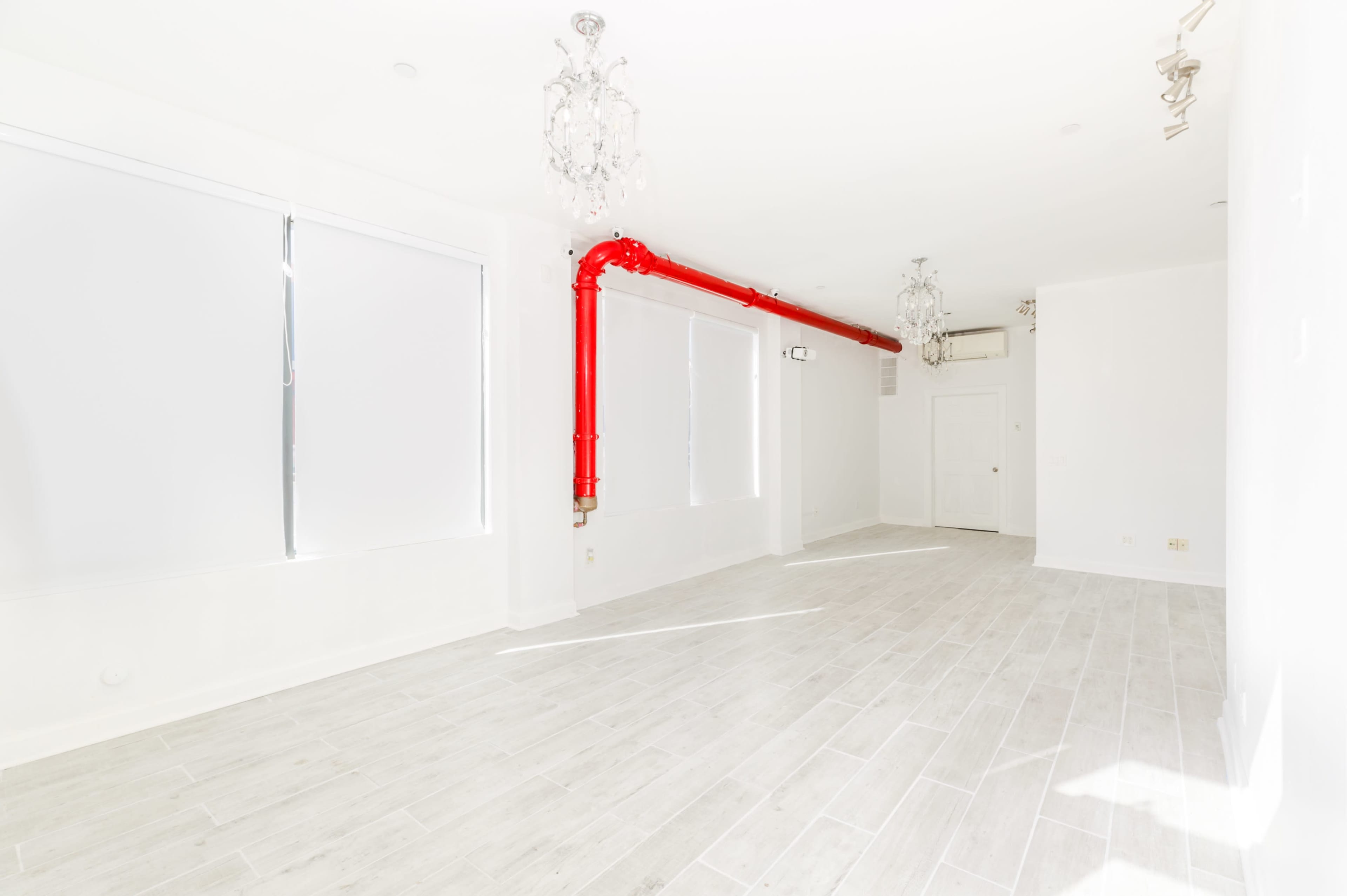 A spacious, empty room with light-colored flooring, large windows with covered blinds, and a prominent red pipe running along the ceiling.