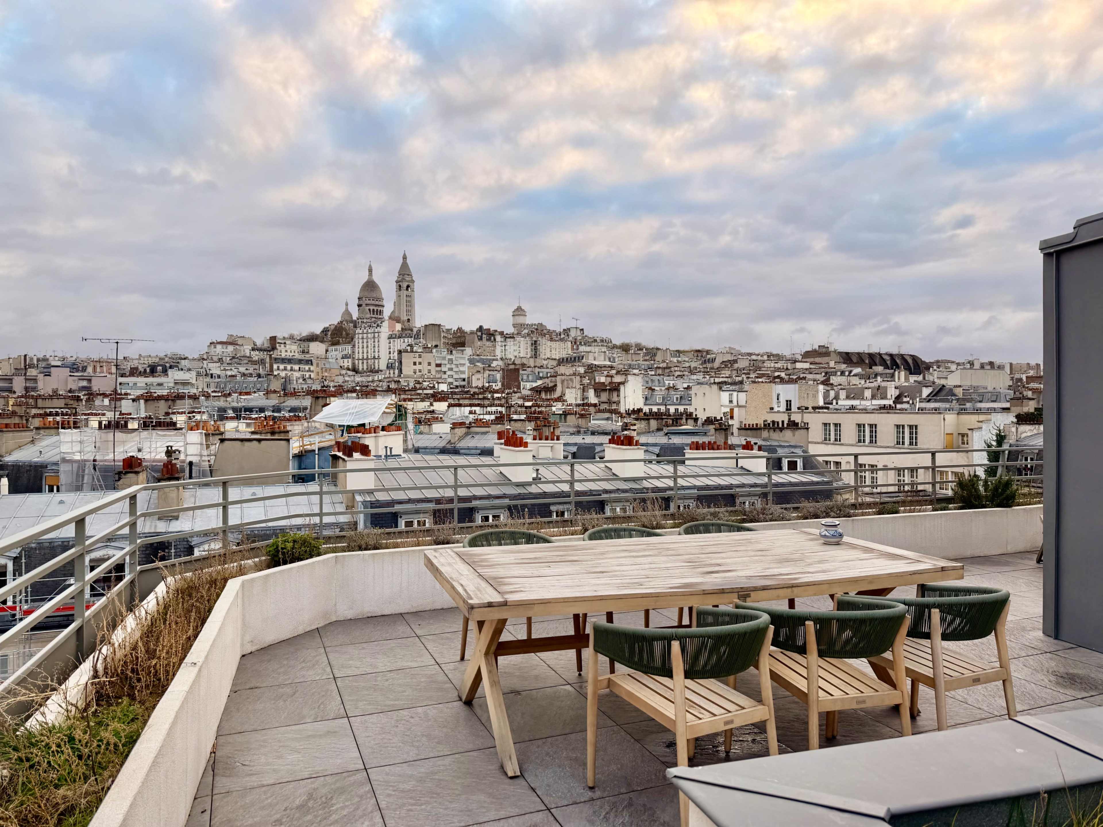A rooftop terrace features a wooden dining table and chairs, overlooking a cityscape with prominent buildings in the distance under a cloudy sky.