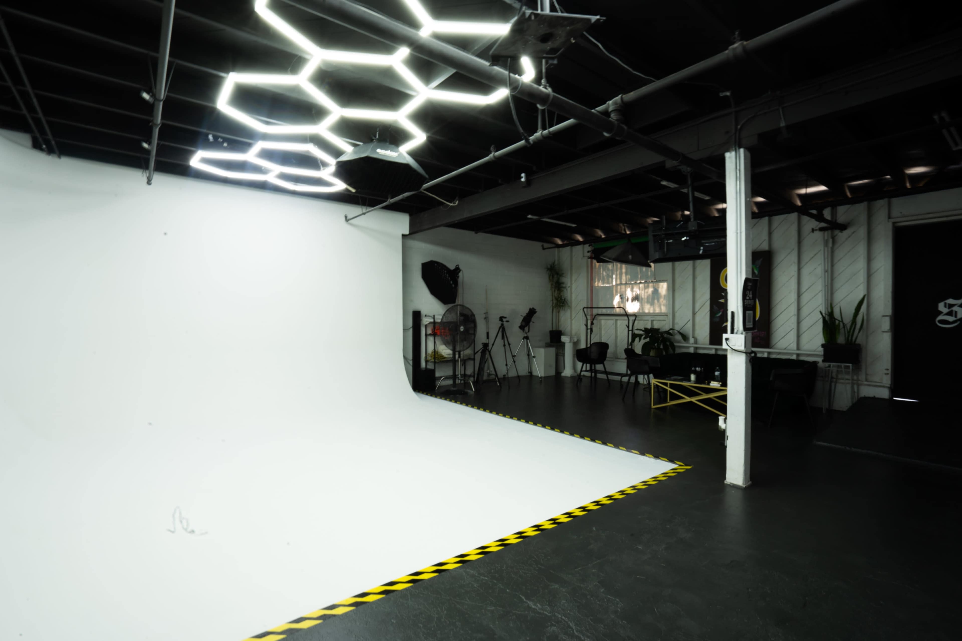 A photography studio features a large white backdrop, hexagonal LED lights overhead, and various equipment arranged along the walls.