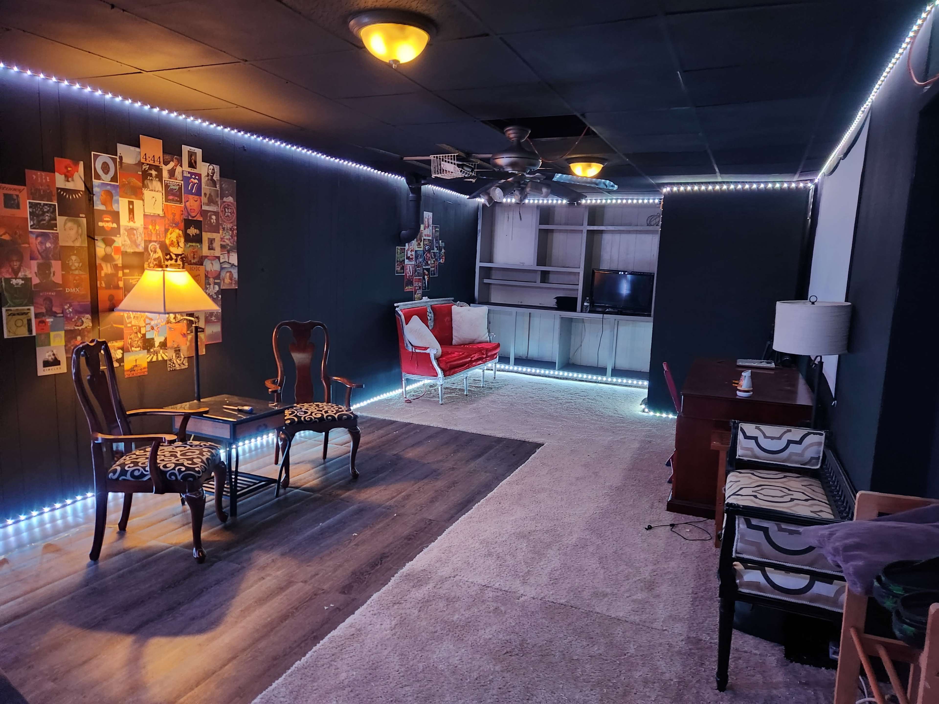 The image shows a dimly lit room featuring a seating area with two chairs and a table, a lamp, a wall display of framed photos, and a workspace with a desk and chair.