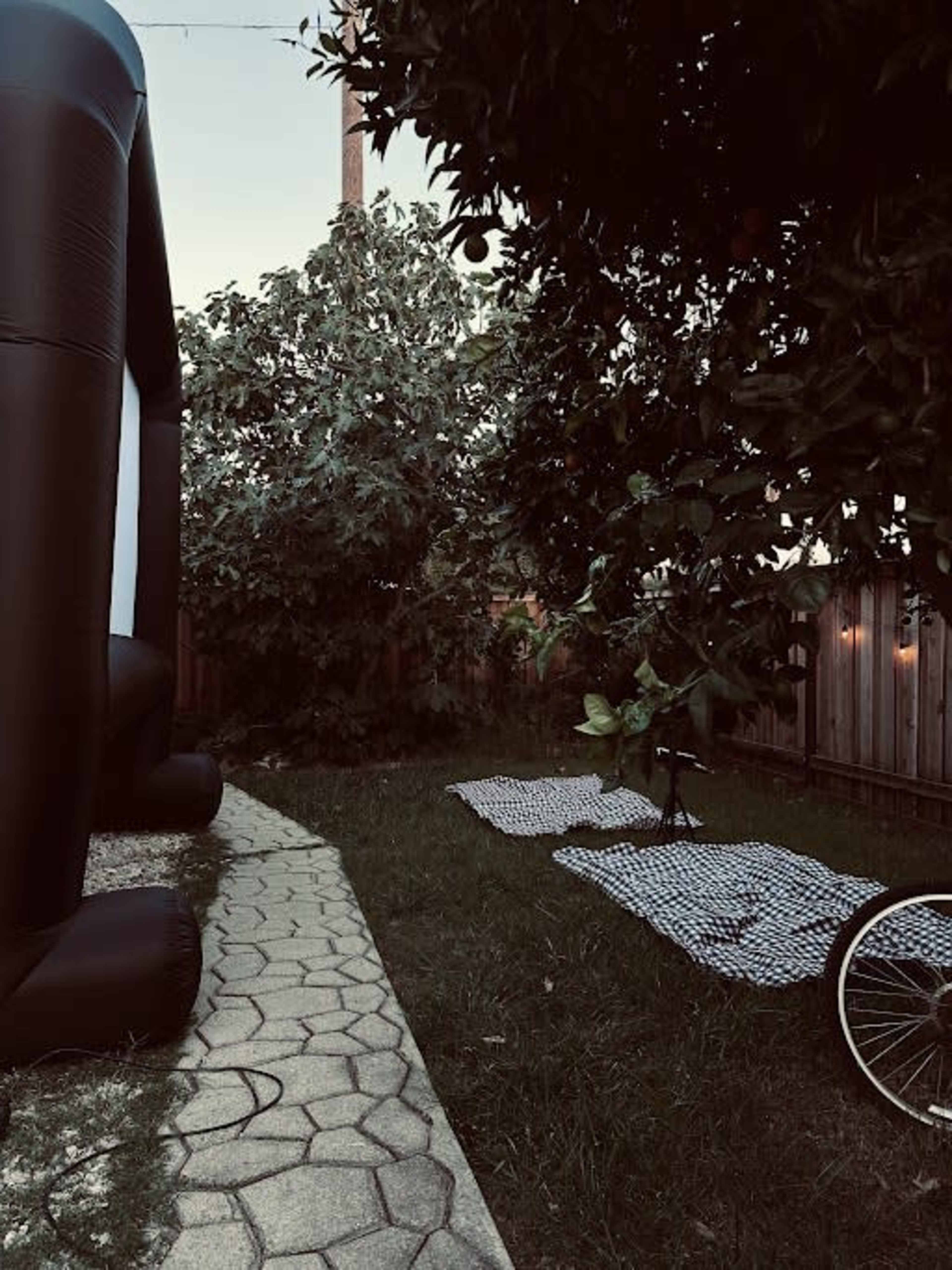 A backyard with a pathway lined by stones, a large inflatable structure to the left, and two checkered blankets spread on the grass.