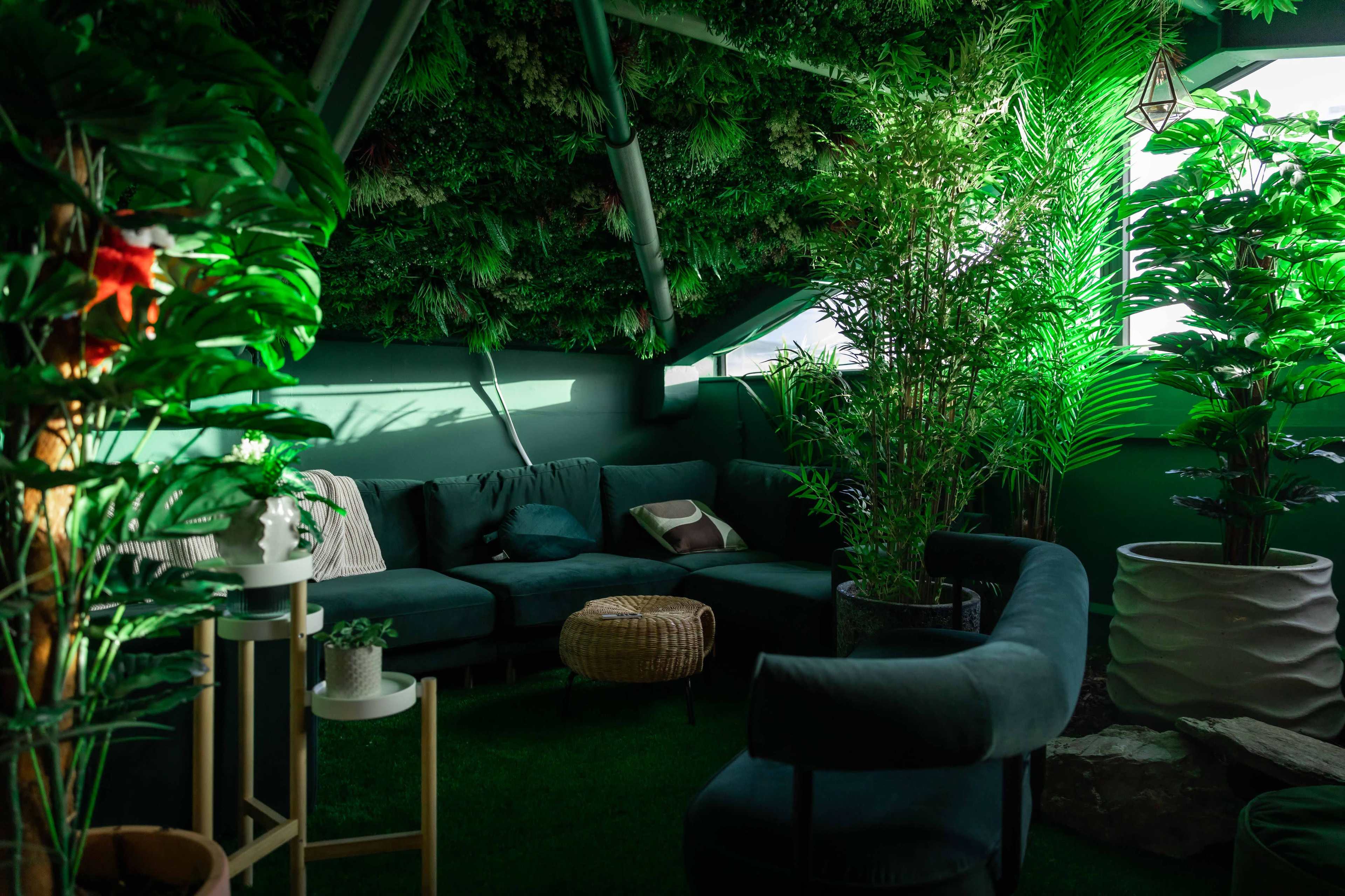 A cozy indoor space filled with various plants, featuring a sectional sofa, a small coffee table, and large windows.