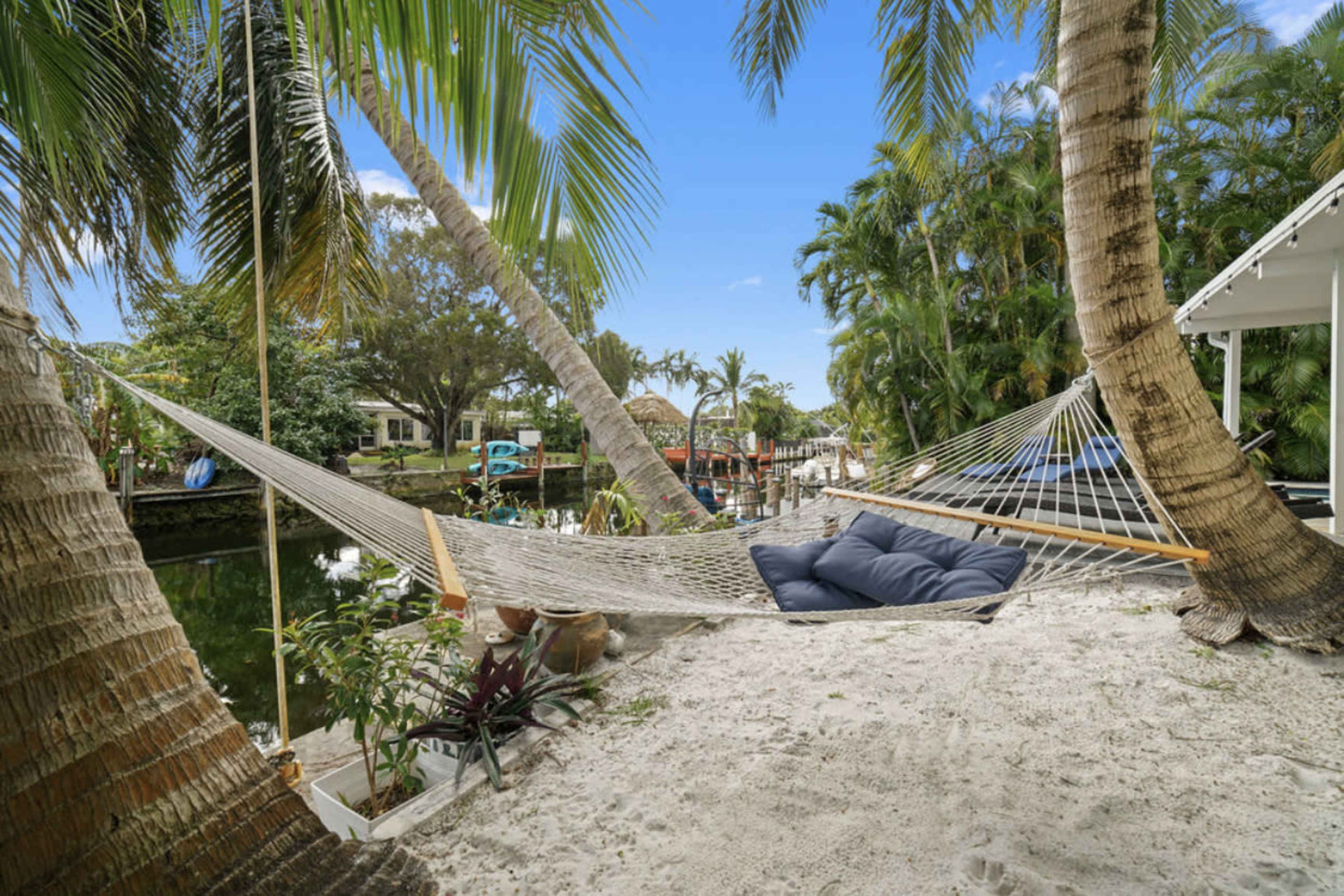 A hammock is suspended between two palm trees on a sandy area beside a calm canal, with boats and greenery visible in the background.