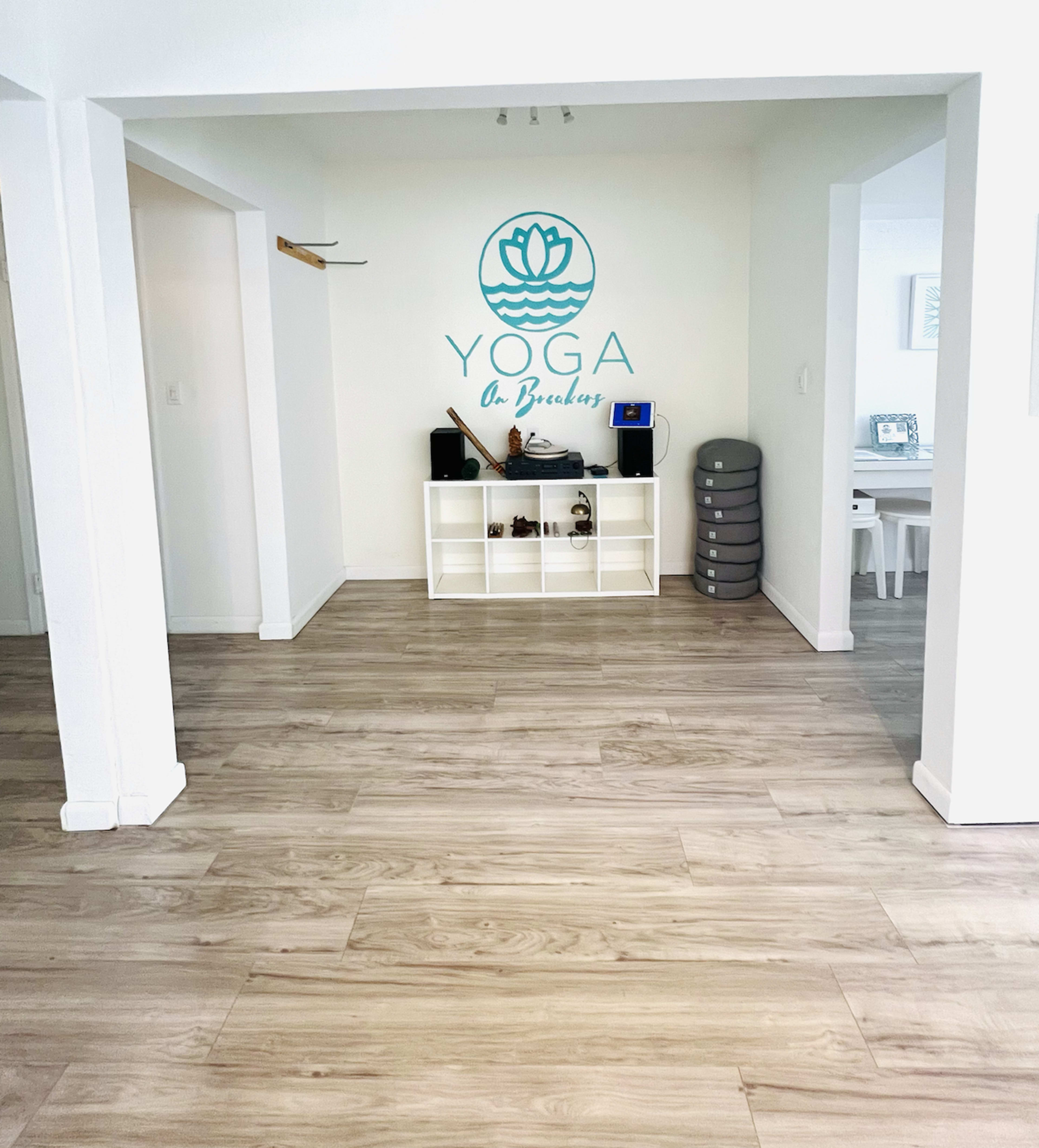 The image depicts a spacious yoga studio with light-colored wooden flooring, a wall featuring a blue lotus logo and the word "YOGA," and a shelf containing speakers and equipment.