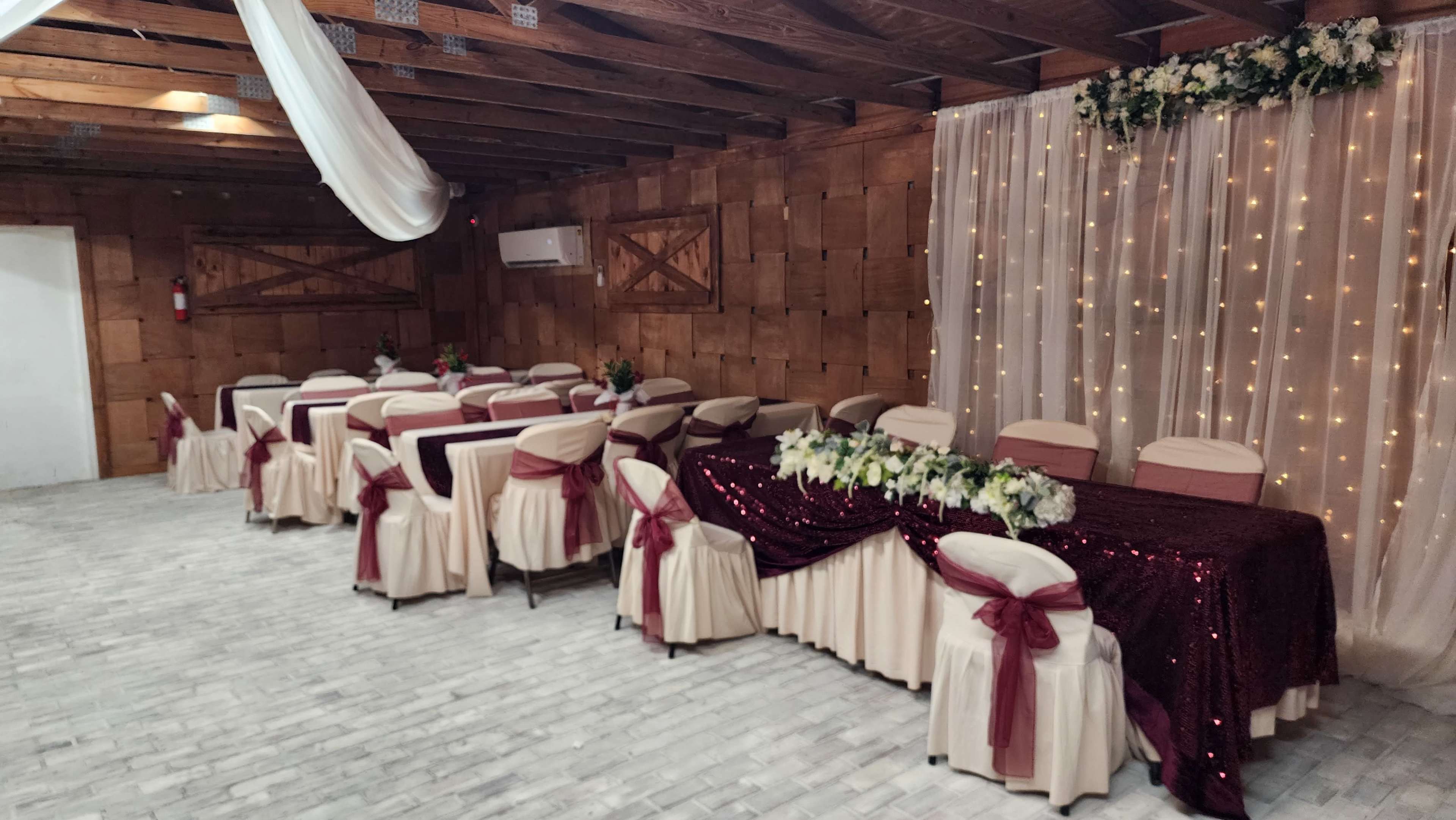 A decorated indoor space with several tables arranged for an event, featuring white tablecloths with maroon sashes and a backdrop of draped fabric with decorative lights and floral arrangements.