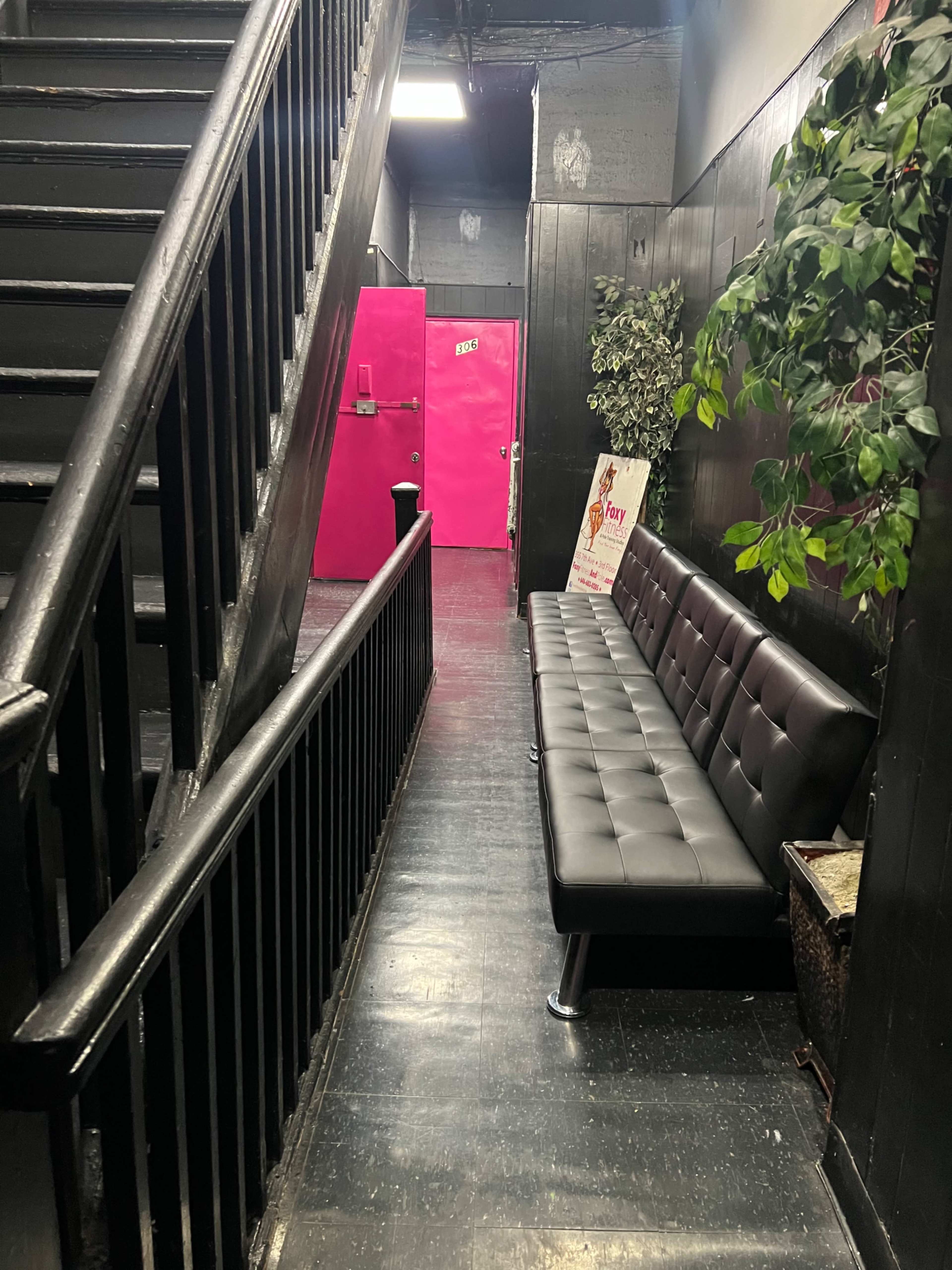 A narrow hallway with a black staircase on one side, a long black bench along the wall, and a bright pink door at the end.