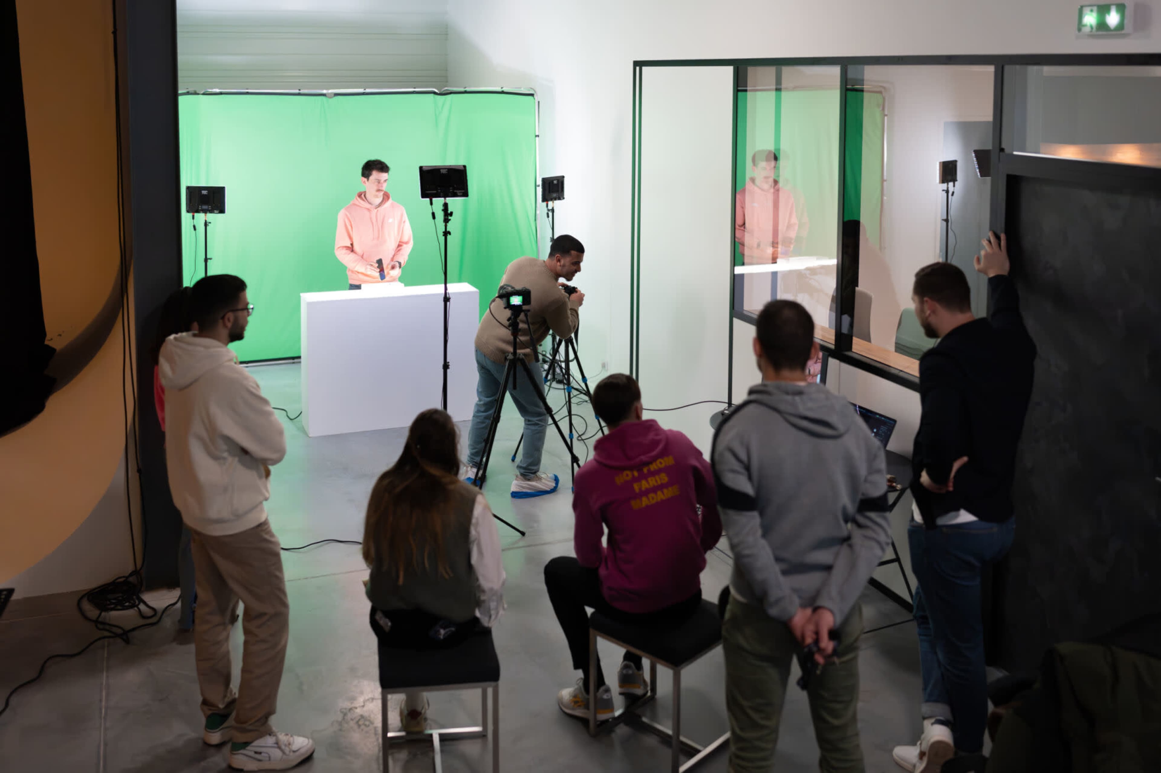 A group of people watches a person presenting in front of a green screen while another individual operates a camera.