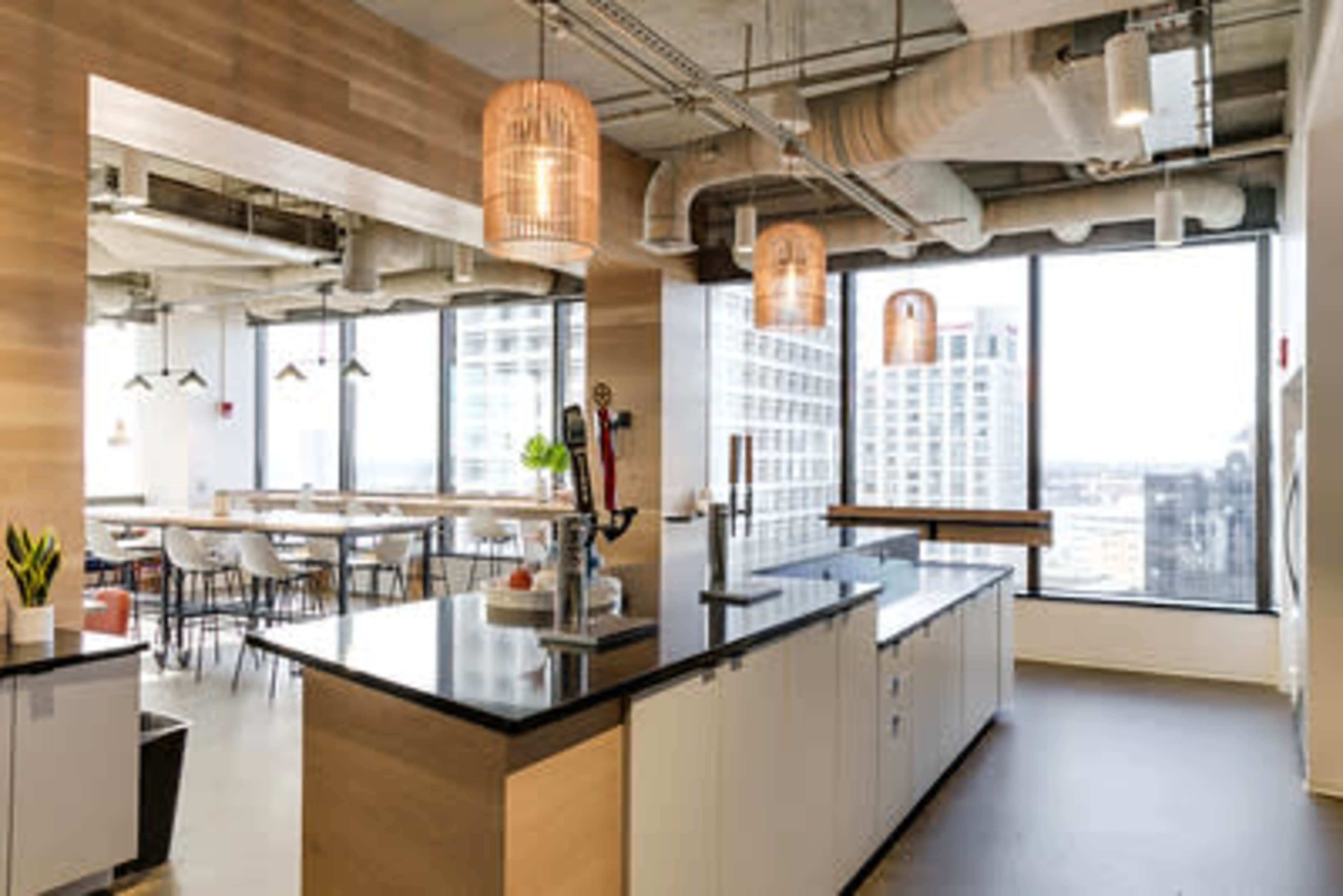 The image shows a modern office kitchen area with a central island, tall windows offering a city view, and pendant lighting hanging from the ceiling.
