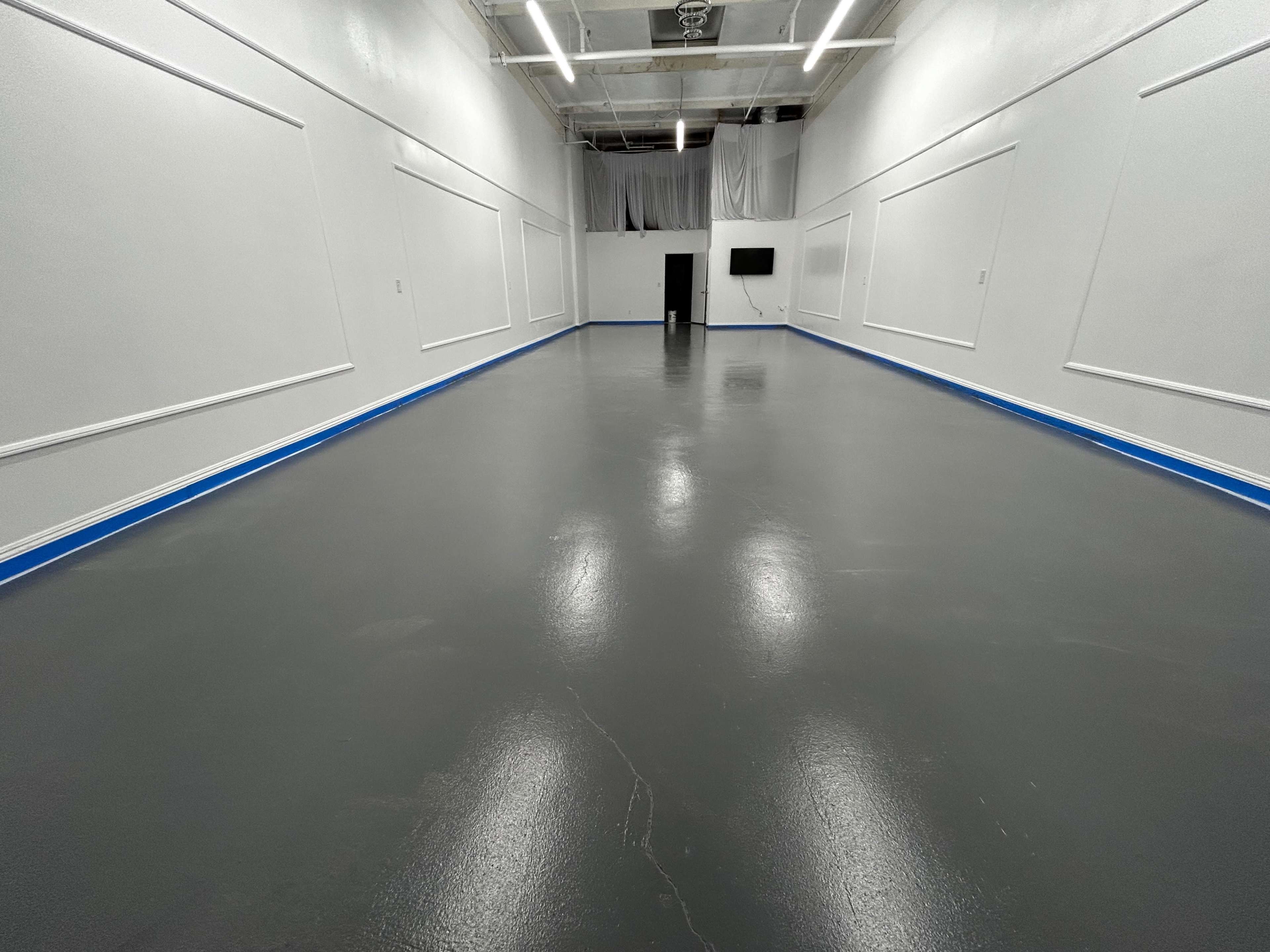 A spacious, empty room with a polished gray floor and white walls, featuring blue trim and a television mounted on one wall.