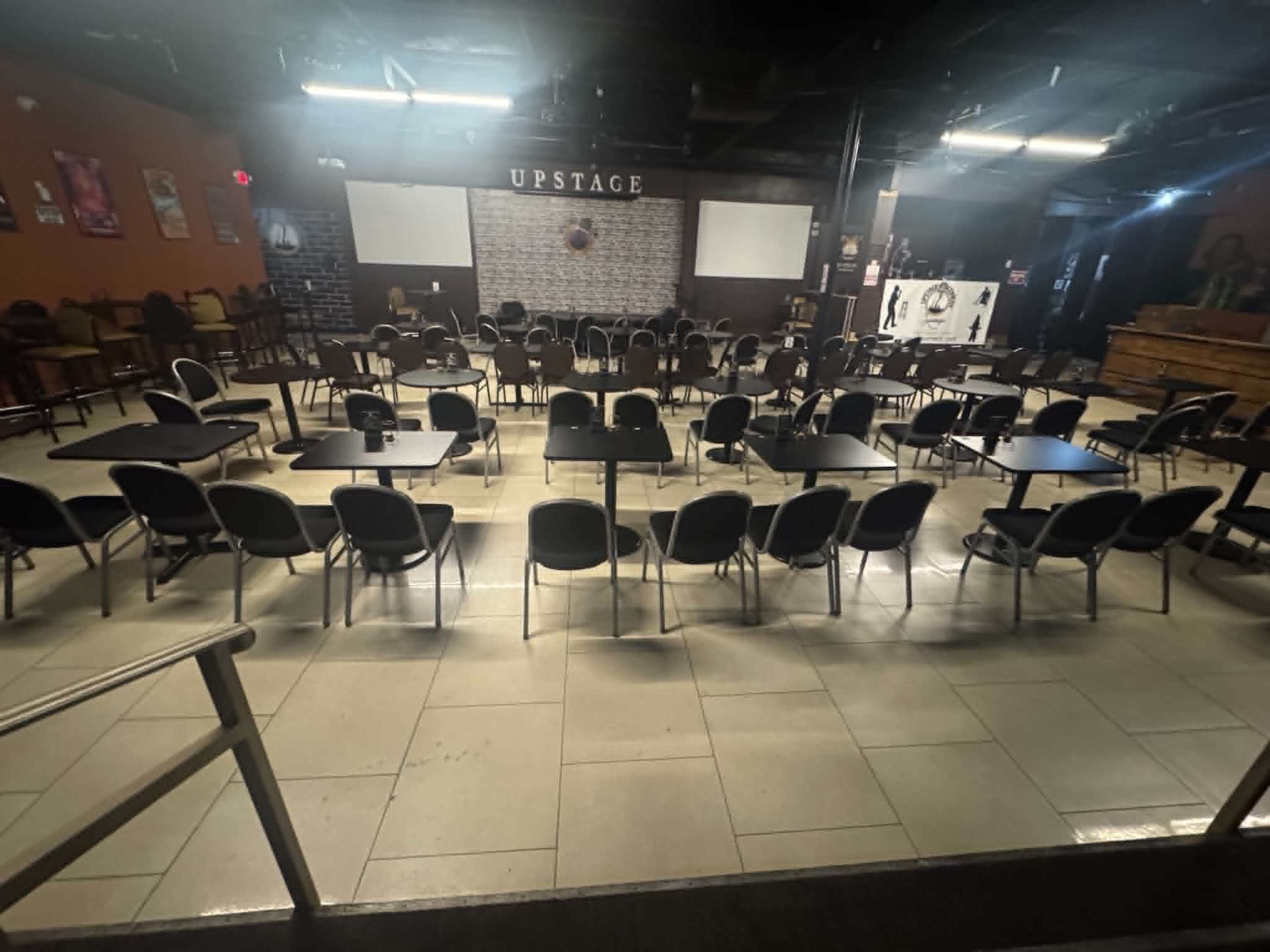 The image shows an empty performance venue with rows of tables and chairs arranged in a rectangular layout, illuminated by overhead lights.