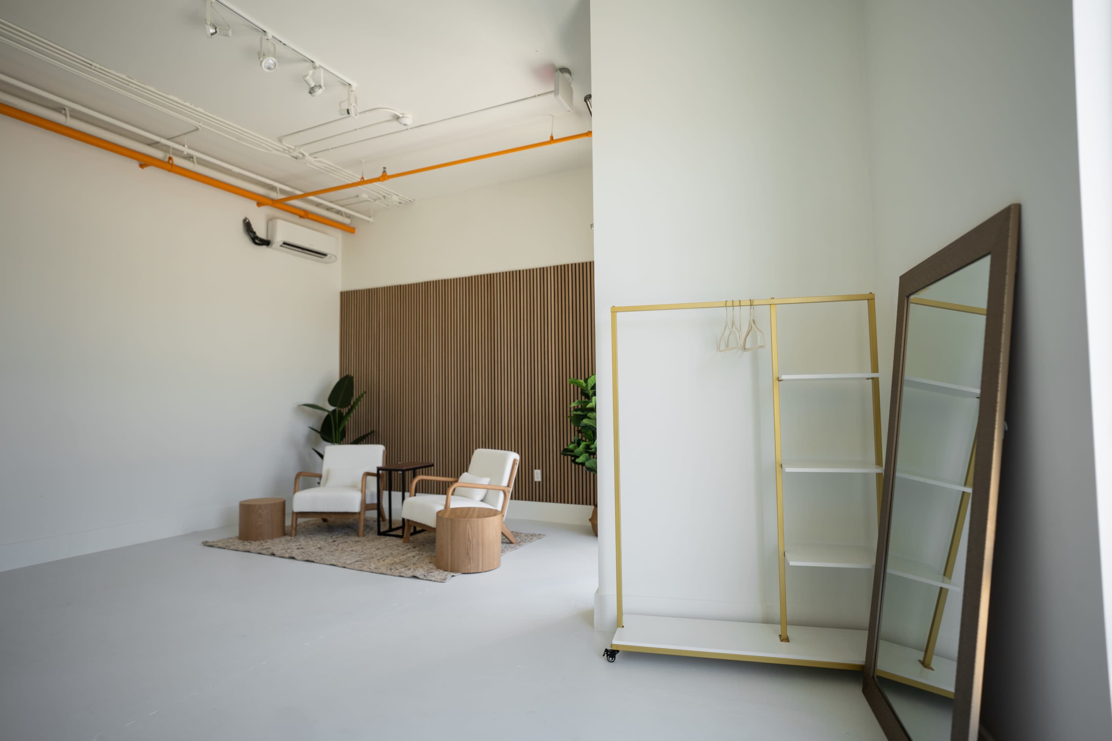 A minimalistic interior space featuring white walls, a wooden accent wall, two chairs, a small table, a large mirror, and a rolling garment rack with hangers.