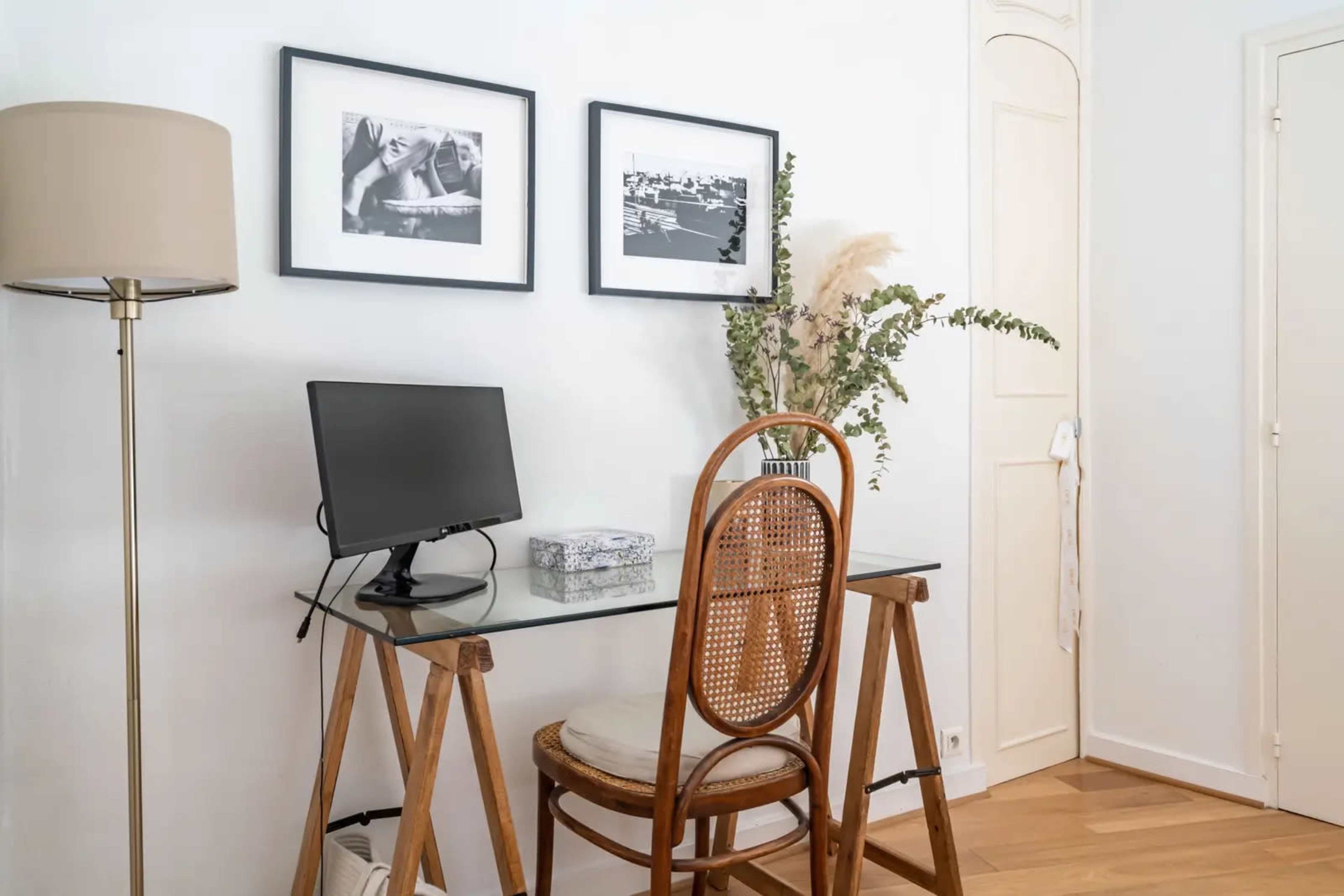 The image shows a minimalist workspace featuring a glass desk, a computer monitor, a wooden chair, and framed photographs on the wall.