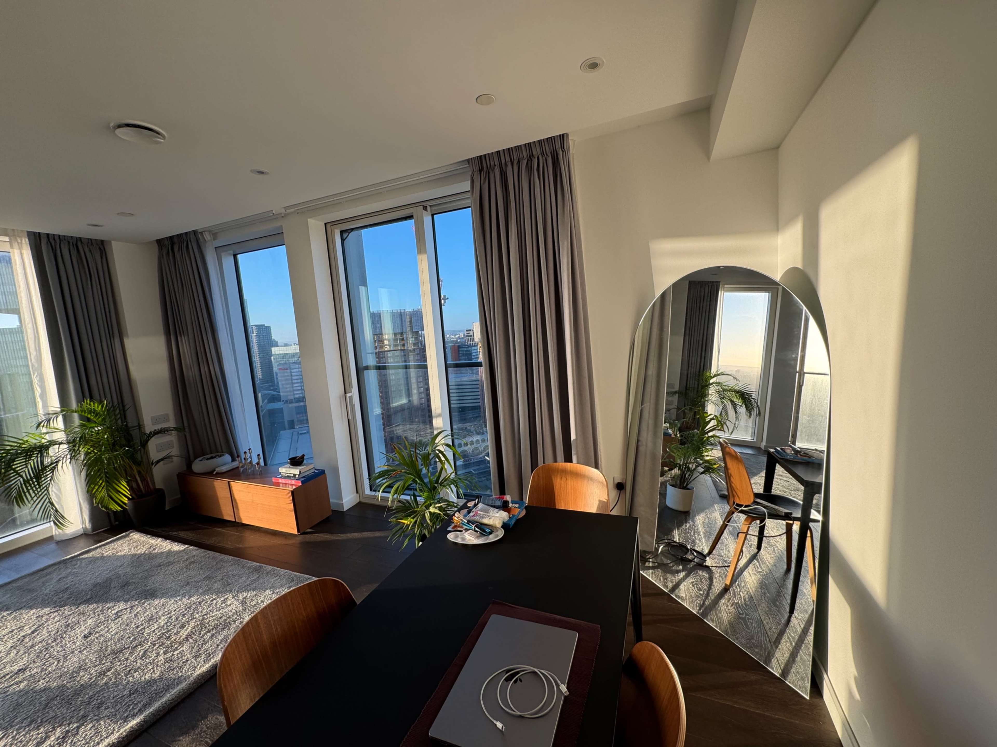 A modern dining area features a black table with wooden chairs, floor-to-ceiling windows, and a large mirror reflecting the cityscape.