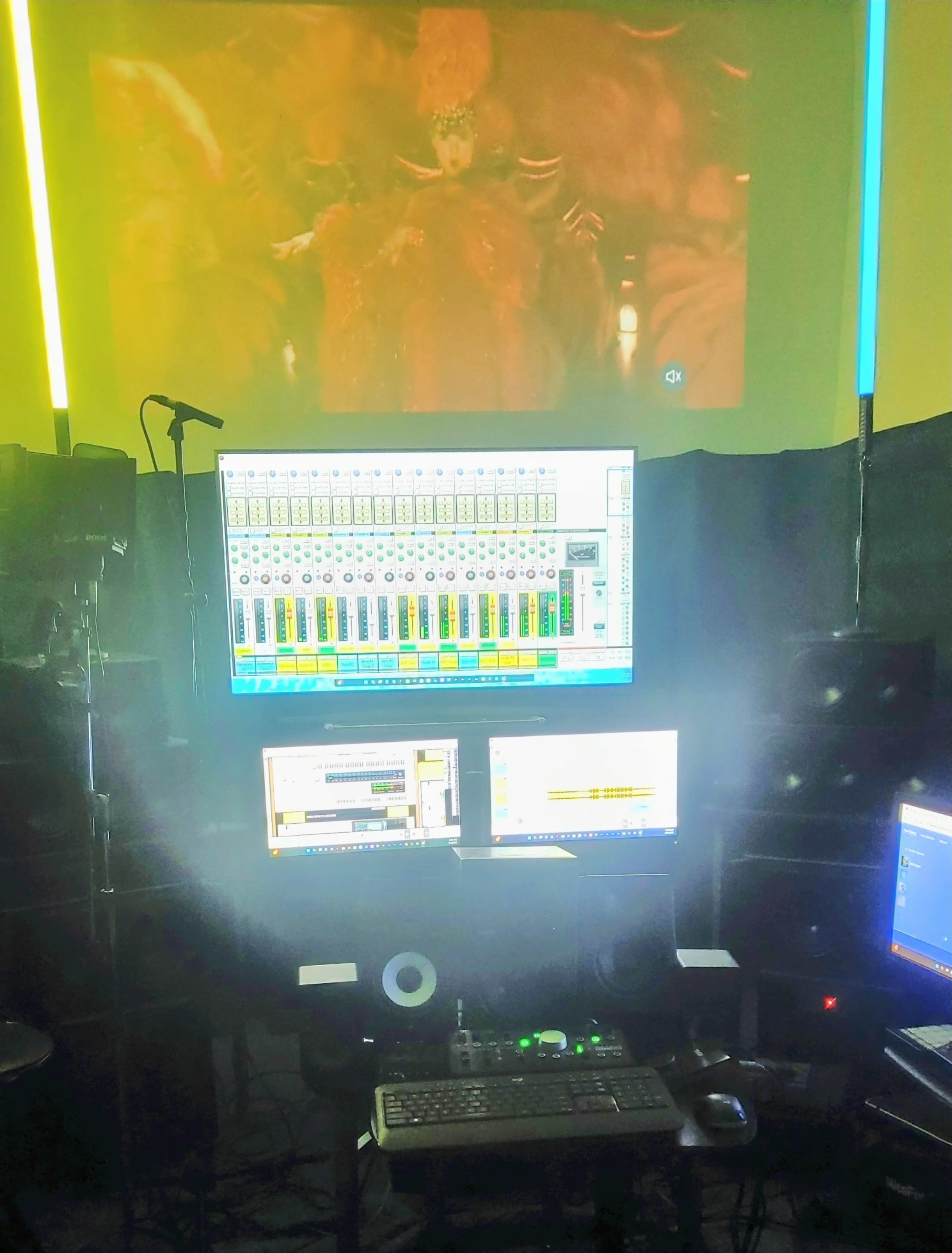 The image shows a music studio with multiple computer screens displaying audio editing software, a microphone, and large speakers, all illuminated by colored lights.